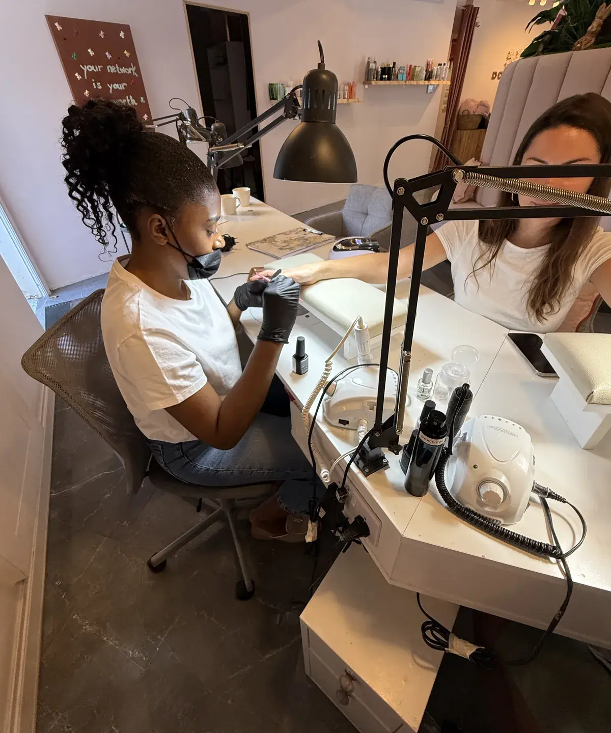A student in a face mask practising Russian manicure with an e-file on a live model at a nail workstation at Nina Georgi Academy