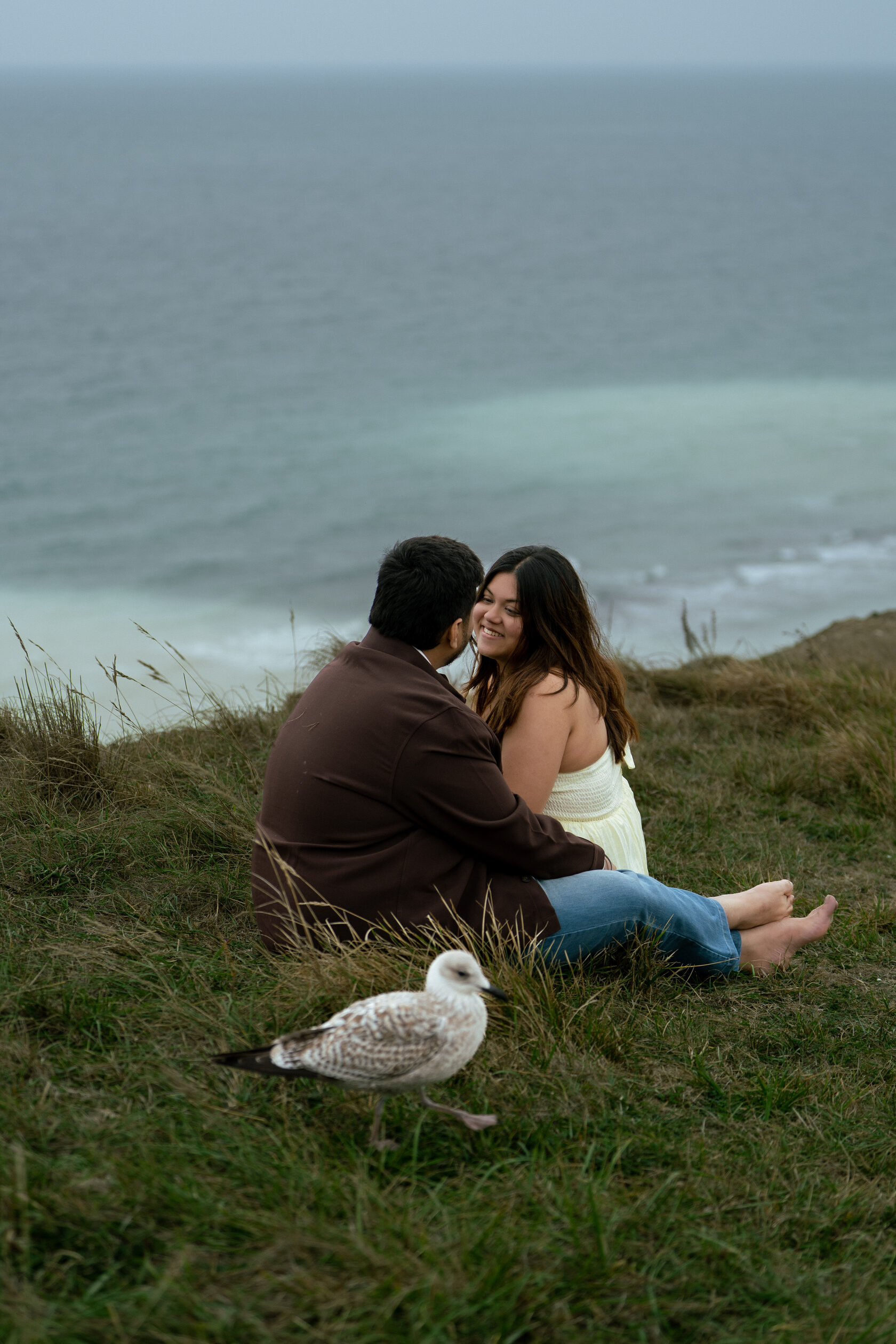 Couple seated together on the grass with the sea behind them