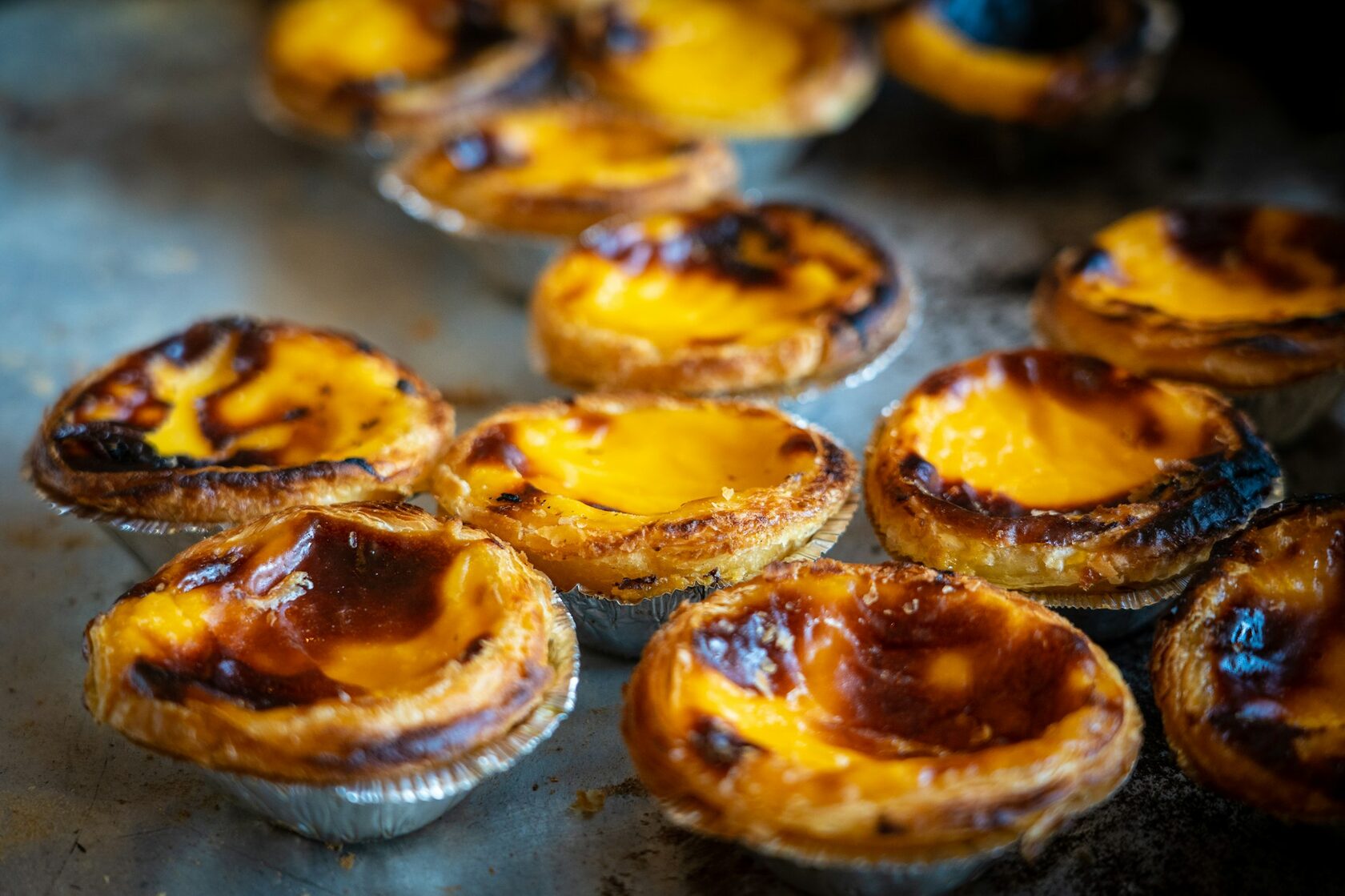 A baker's tray of golden custard tarts fresh from the oven, a cinnamon shaker beside them