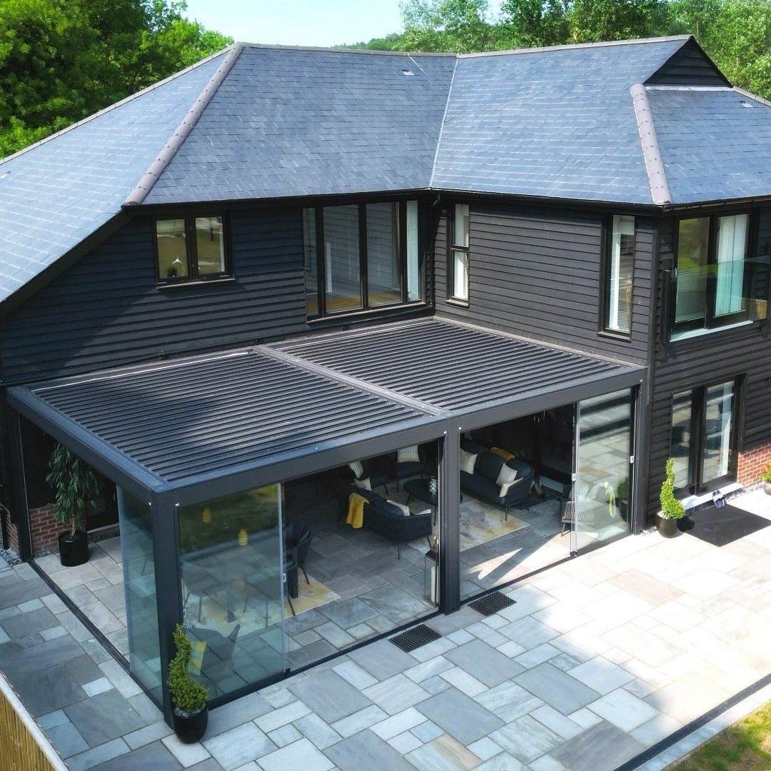 Dark timber house with a louvered pergola and glass walls over a stone terrace