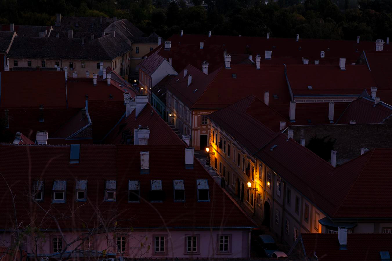 Aerial dusk view of a historic European town with red rooftops and warmly lit windows.