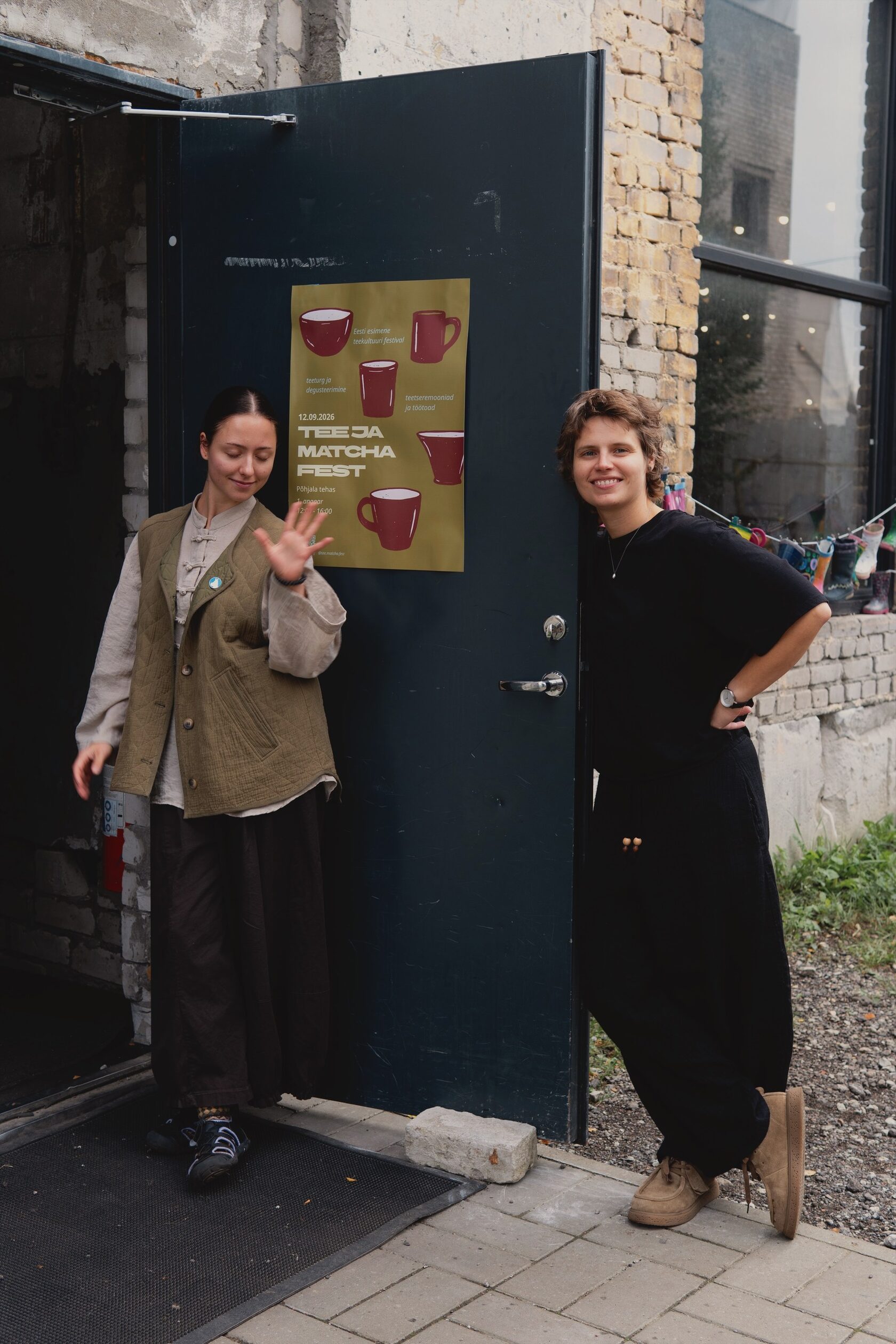 Tanhel and Emilija by an open factory door with a Tea & Matcha Fest poster