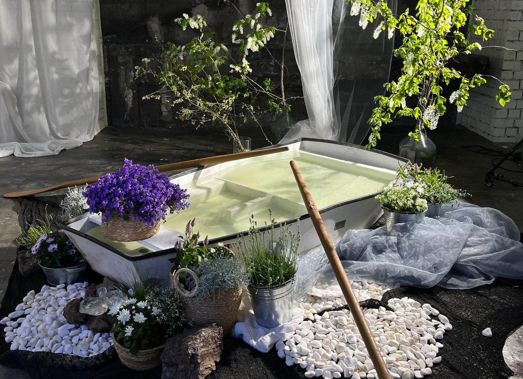 A white rowing boat filled with water, surrounded by potted flowers, white pebbles and blossoming branches — a set at NOT A STUDIO