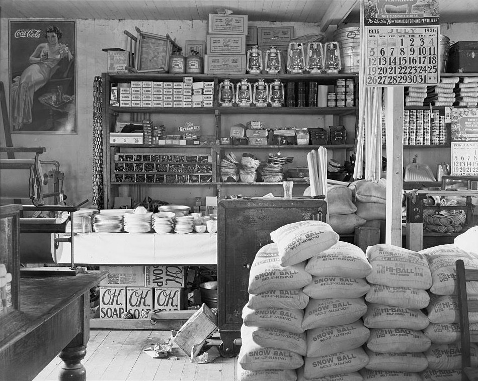 Archive photograph of a 1936 general store: shelves of boxes, lanterns and dishes, with stacked flour sacks in front