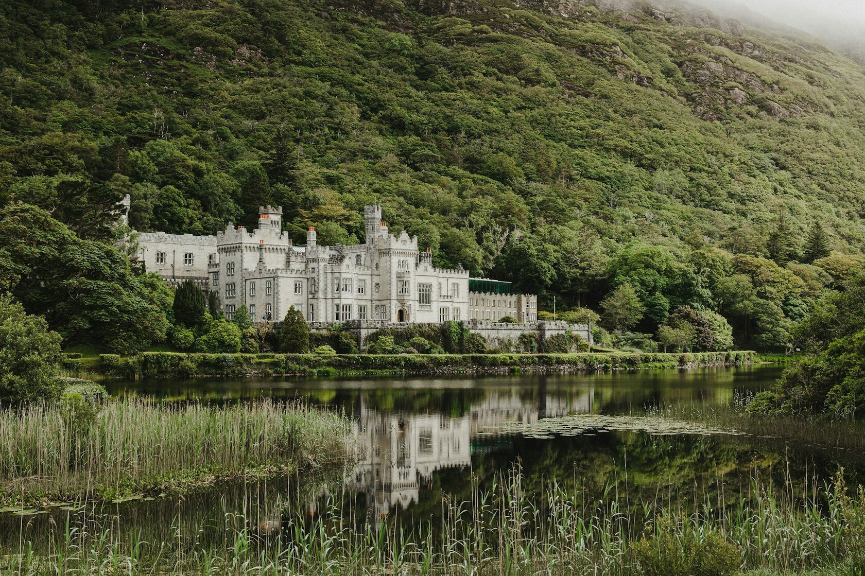 Kylemore Abbey in Connemara, County Galway, its Gothic façade mirrored in the still lake beneath wooded mountains, soft golden light and a little mist on the water.