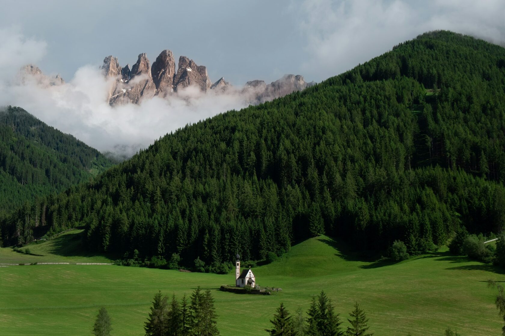 The little white church of St. Johann in Ranui standing alone in a bright green meadow in Val di Funes, with the jagged Odle peaks of the Dolomites rising behind it in warm golden late-afternoon light