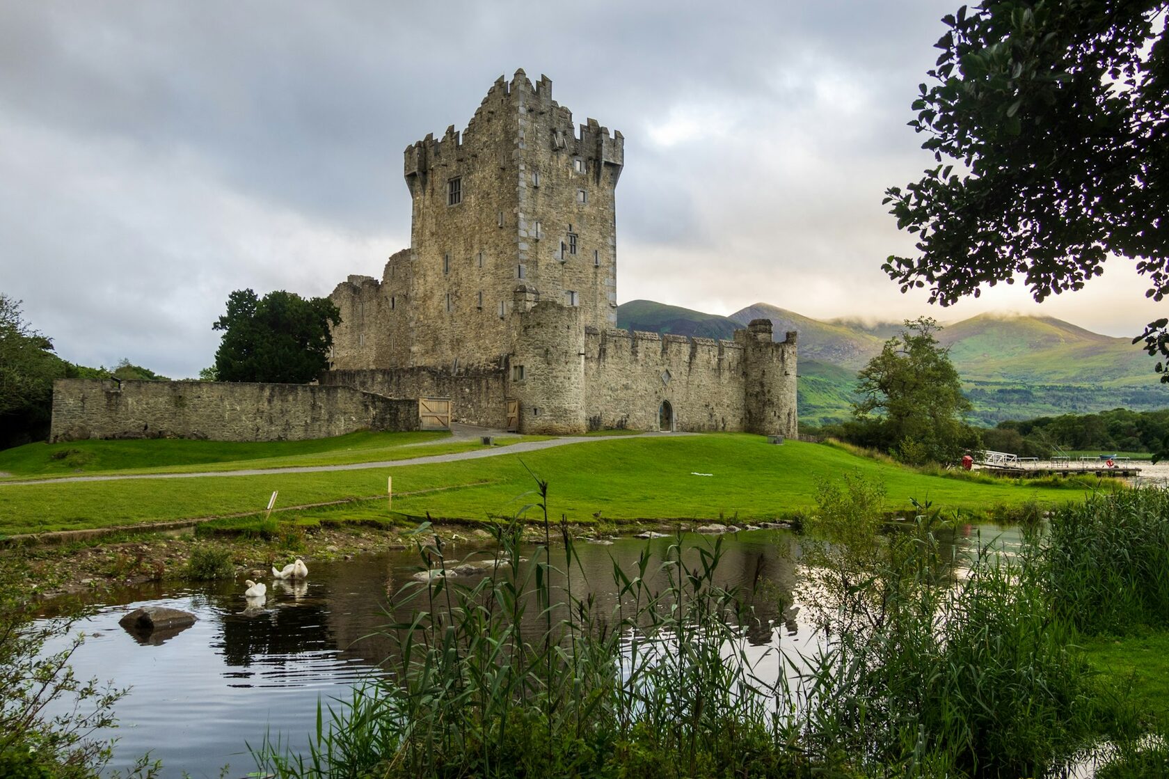 Ross Castle on the shore of Lough Leane in Killarney National Park at sunset, mountains glowing behind and the castle reflected in the calm lake.