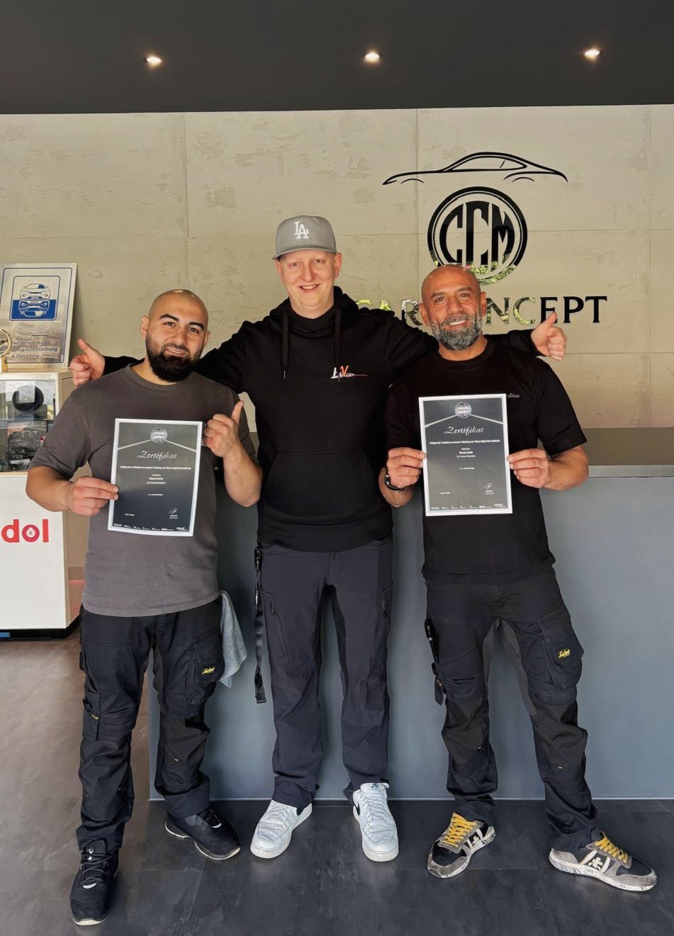 Drei Männer in der Werkstatt der Car Concept Manufaktur präsentieren ihre Scholl-Concepts-Zertifikate. In der Mitte Master Trainer Kevin Vöhringer, flankiert von zwei Teammitgliedern.