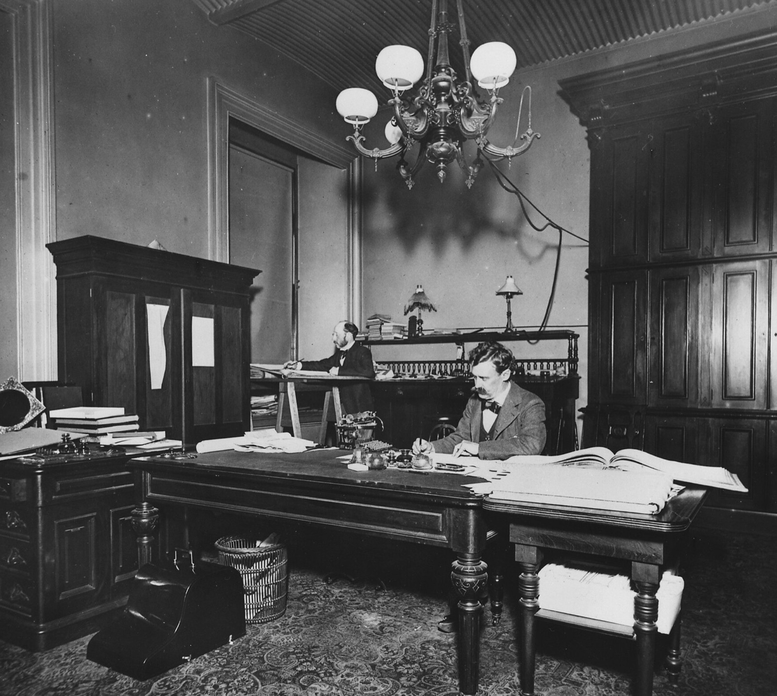 Archive photograph of the San Francisco Mint office: two clerks writing in large account books under a chandelier