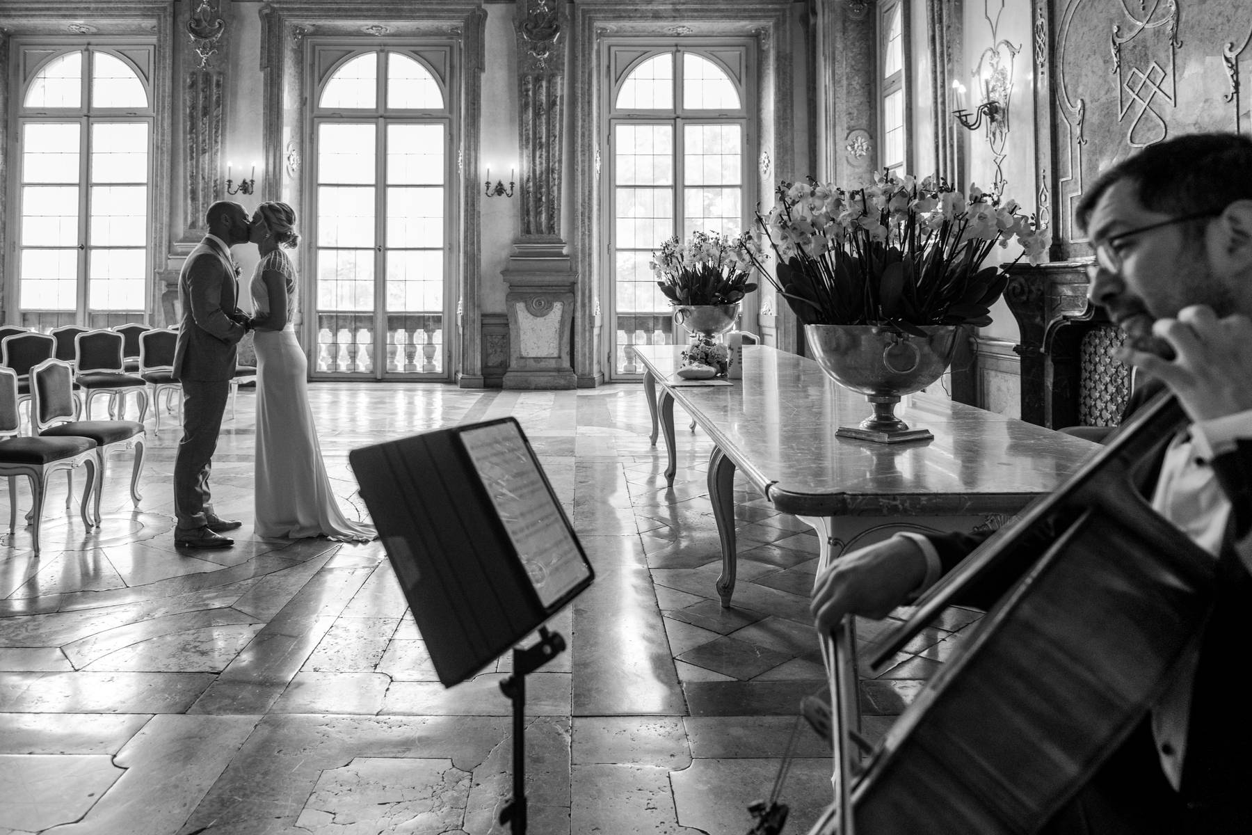 Black-and-white wedding photograph of bride and groom kissing in the Marble Hall at Schloss Mirabell, with a cellist in the foreground