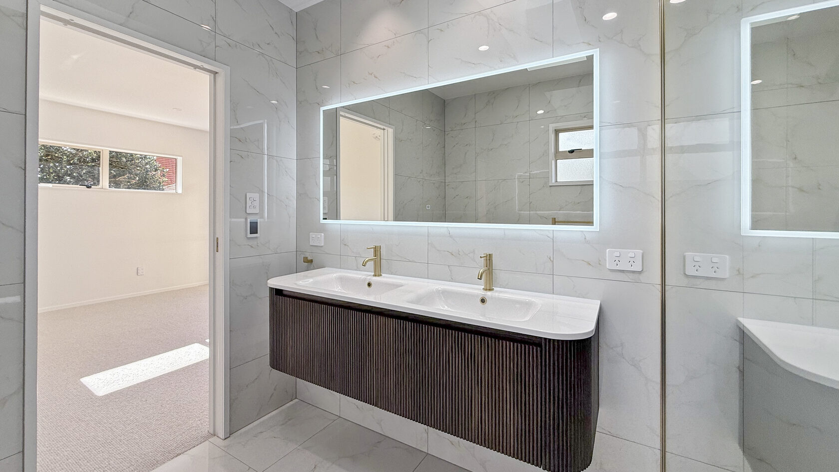 St Heliers bathroom with double vanity, illuminated mirror, marble-look tiles and brass taps.