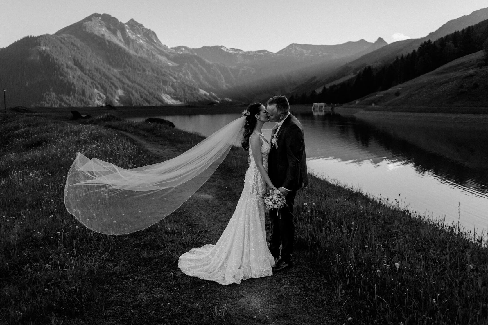 Bride and groom kissing beside a peaceful alpine lake, with the bride's long wedding veil flowing in the wind against a mountain backdrop