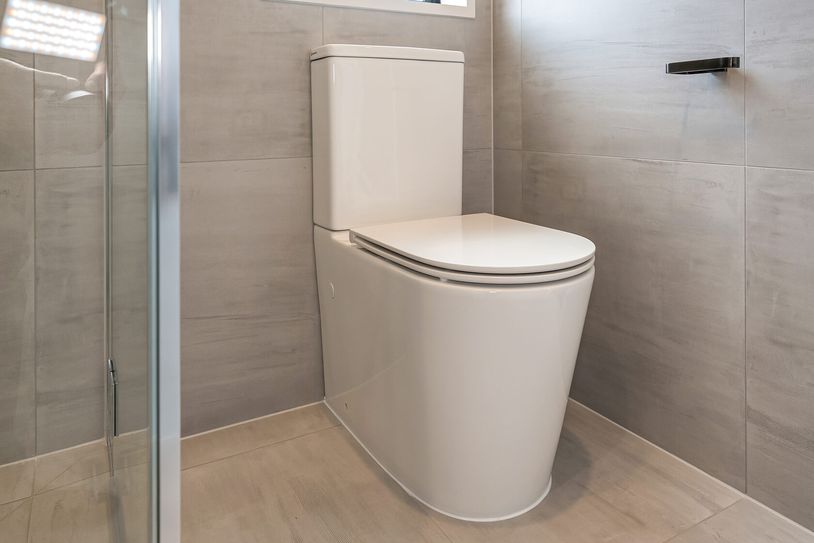 Contemporary close-coupled toilet with a sleek, curved design, set against large-format beige wall tiles and light-toned floor tiles, positioned beside a frameless glass shower panel for a clean, minimalist bathroom finish.