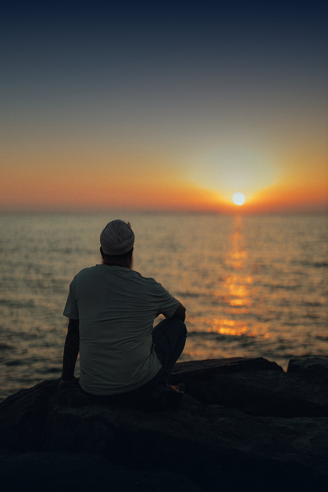 Jugat Guru seated on rocks by the sea at sunset, seen from behind