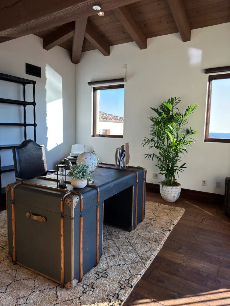 A tall areca palm in a white planter beside ocean-view windows in a coastal home office with a beamed ceiling