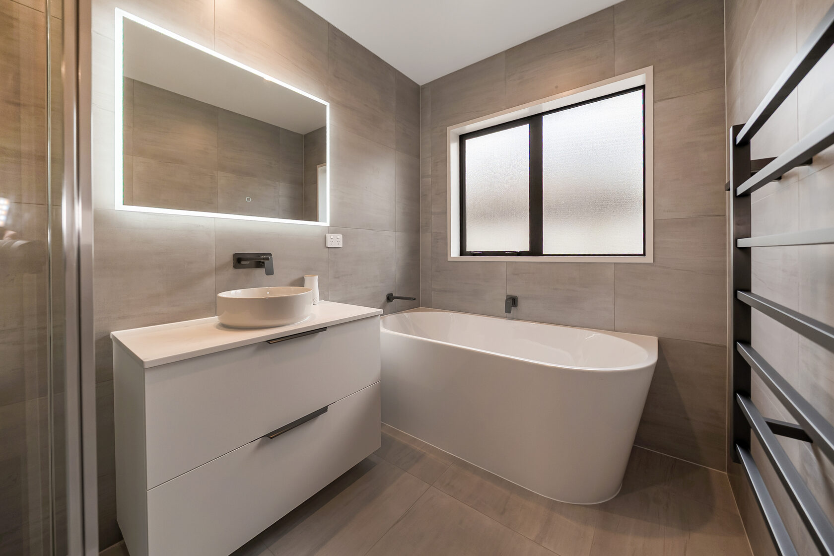 Contemporary bathroom featuring a freestanding bathtub, floating vanity with vessel basin, matte black wall-mounted tapware, LED backlit mirror, large-format stone-look tiles, and a frosted window for natural light and privacy.