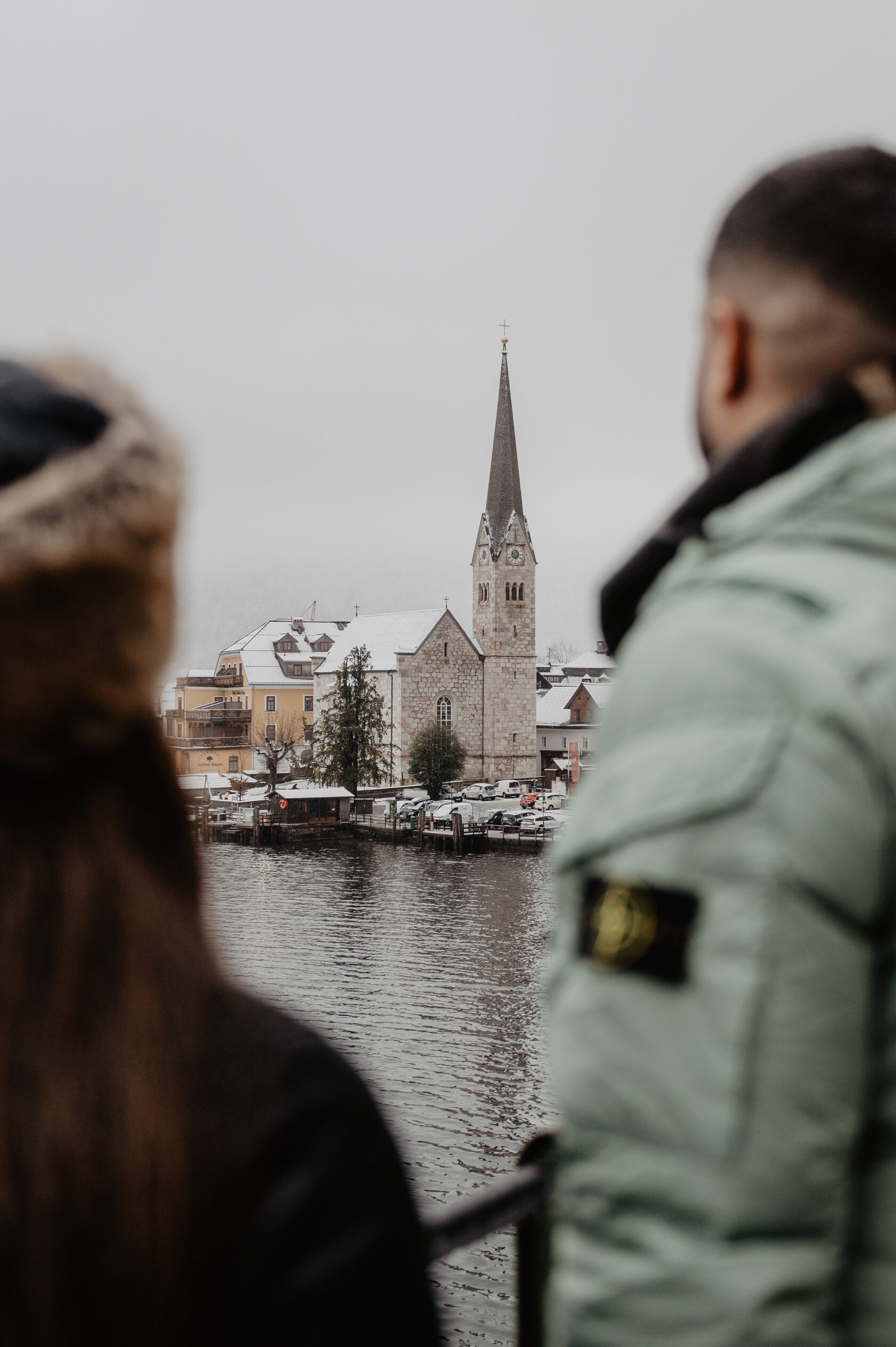 Engaged couple by an alpine lake with a historic church tower and winter village scenery
