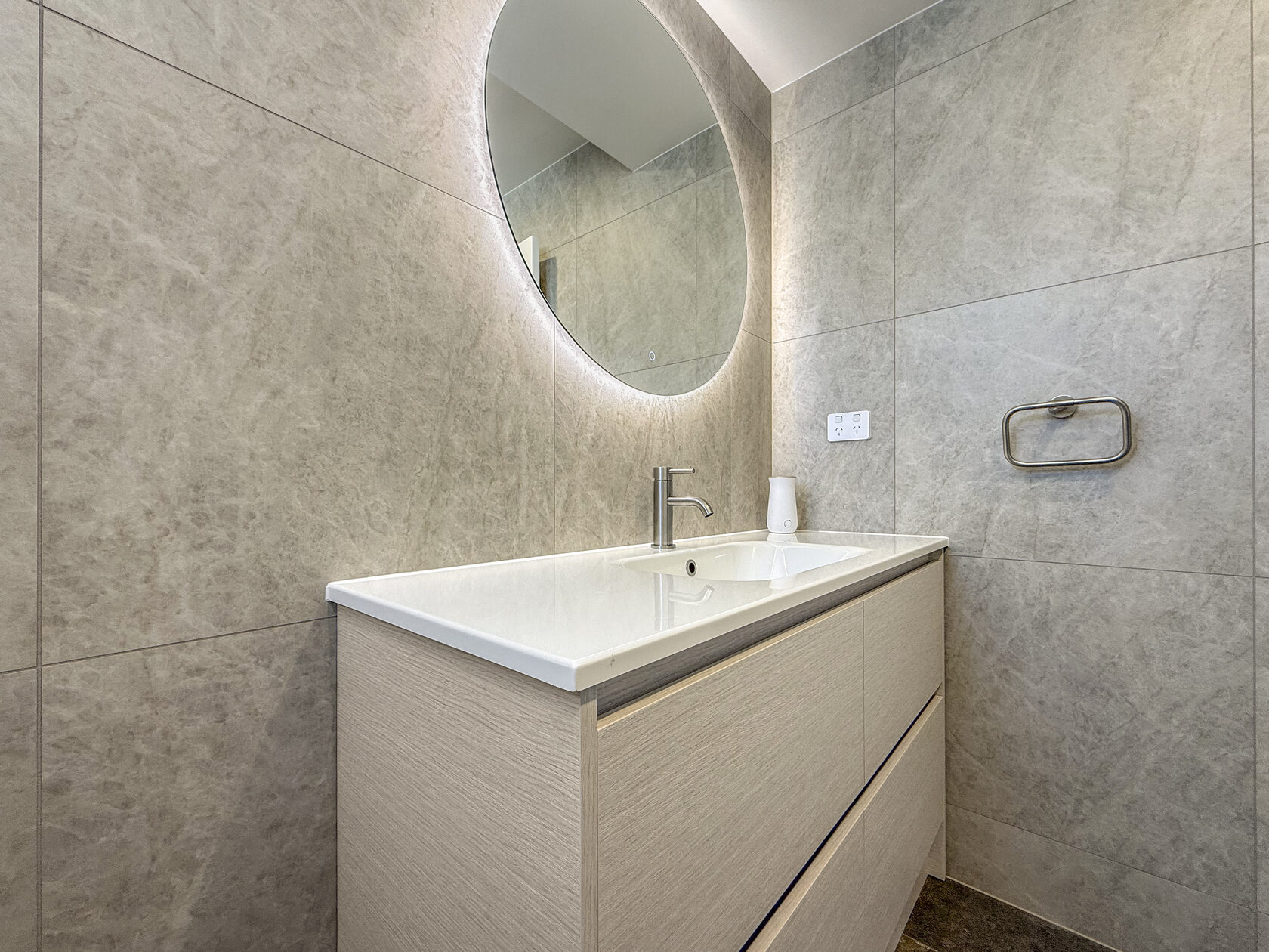 Pale bathroom vanity, round mirror and large grey wall tiles