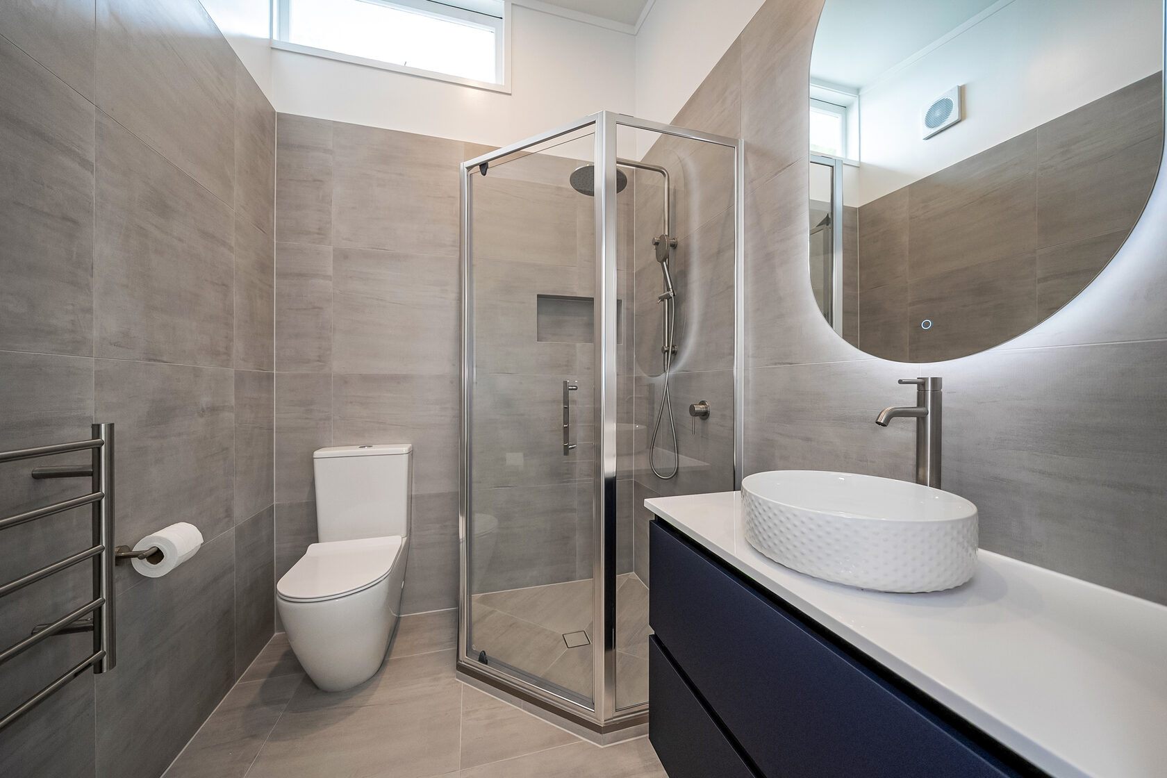 Modern bathroom with glass corner shower, grey tiled walls, floating vanity with vessel sink, and backlit mirror.