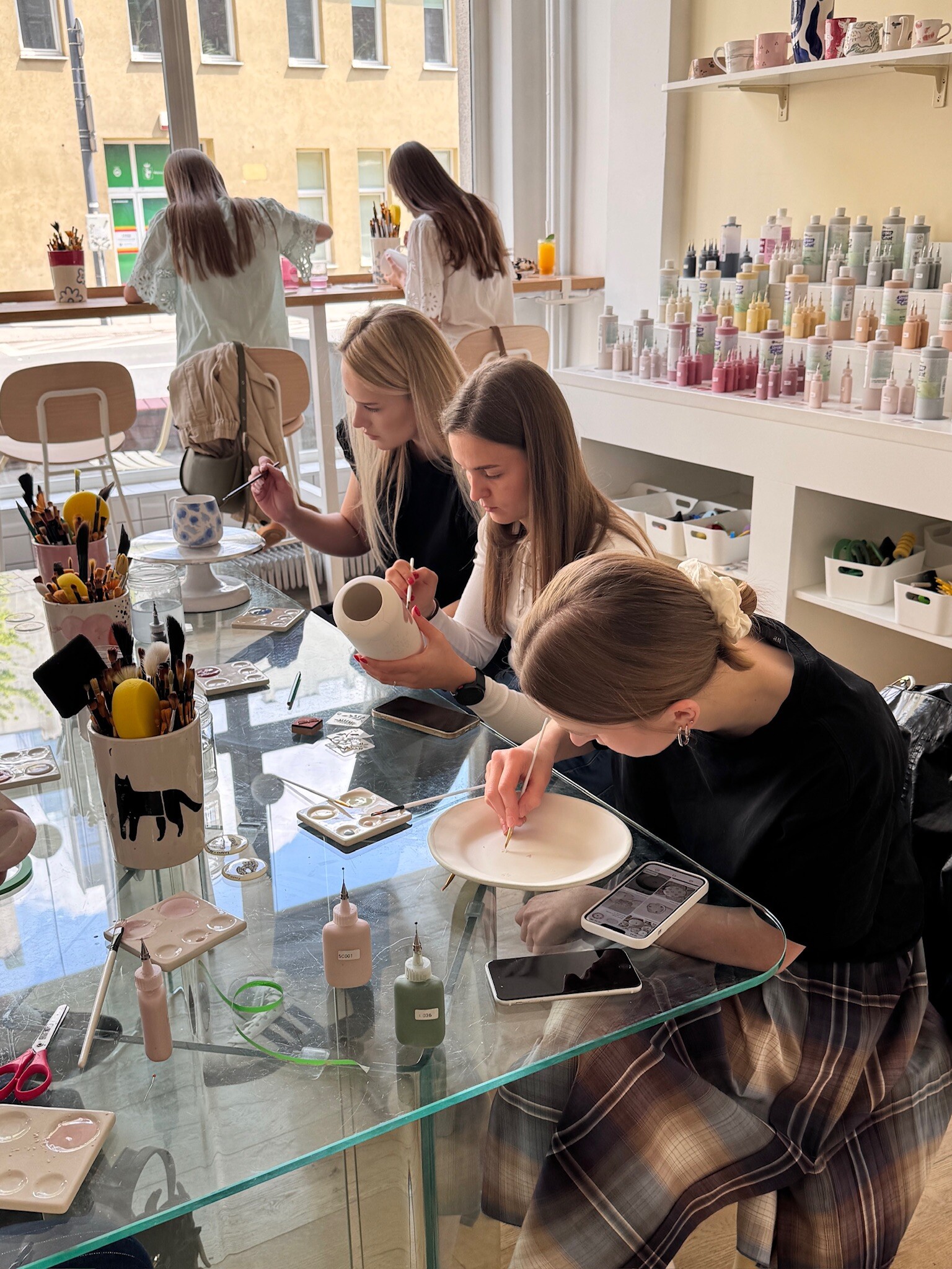 A team painting ceramics at a glass table at Create Cafe in Warsaw — shelves of paints and a yellow tenement house outside the window