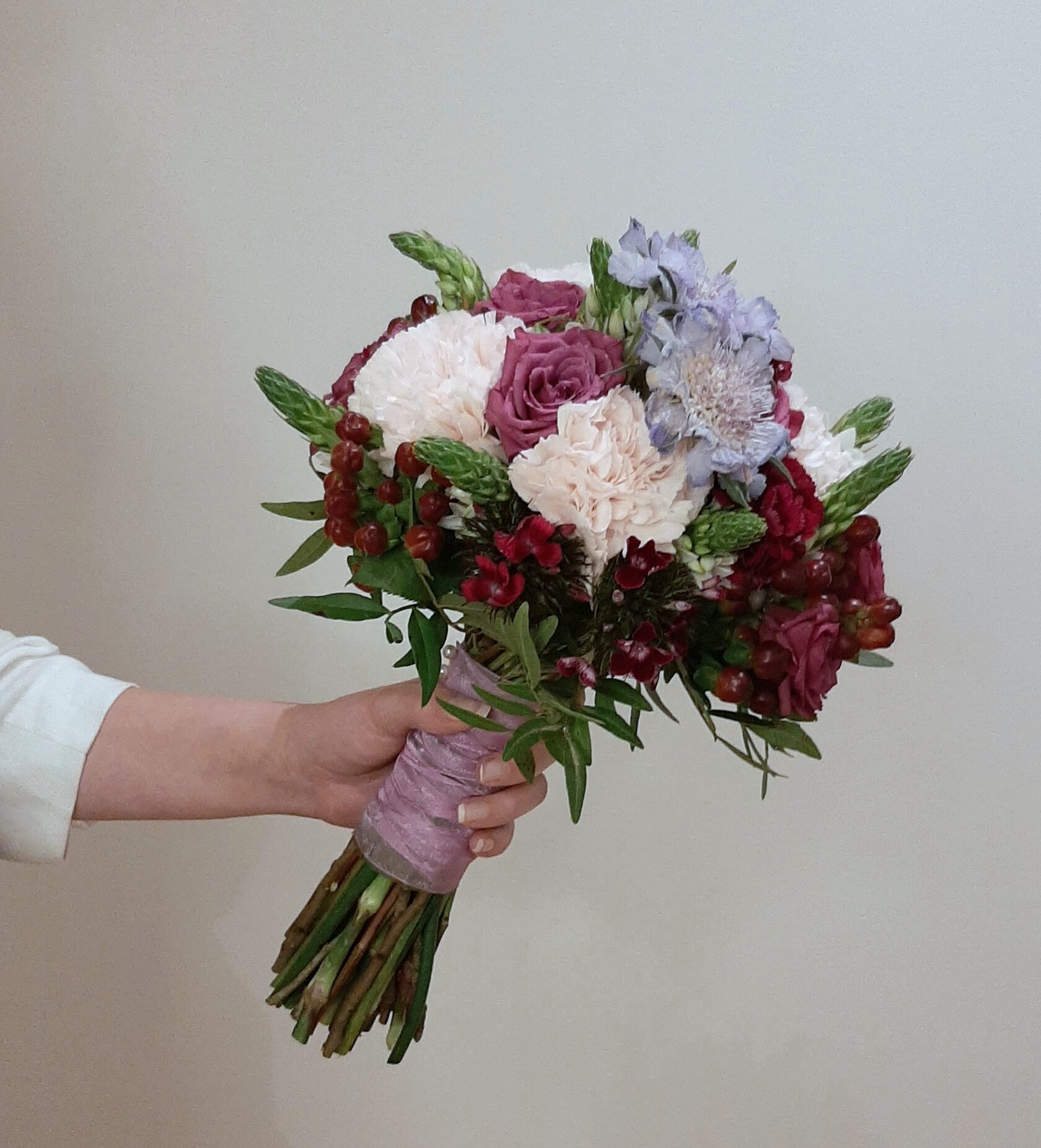 a beautiful bridal bouquet accented with delicate creamy pink peony roses, large white dianthus (carnations), lilac-blue scabiosa and rich burgundy berry accents