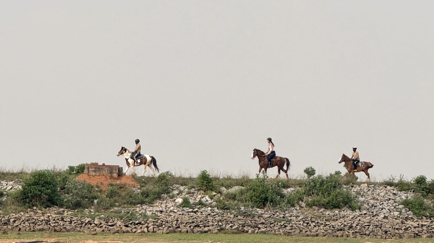 Three riders along the tank bund at Punanai under a haze sky, the stone revetment below.