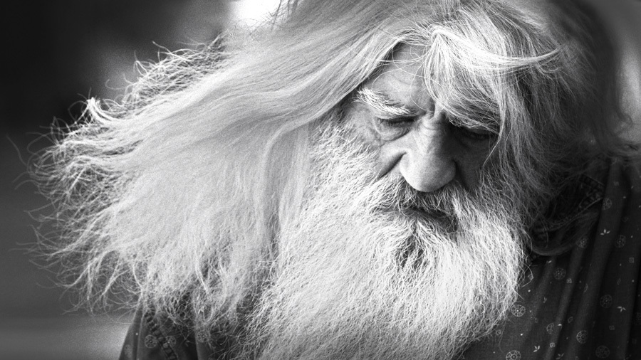 Portrait of an elderly man with long white hair and beard, eyes closed in quiet reflection.