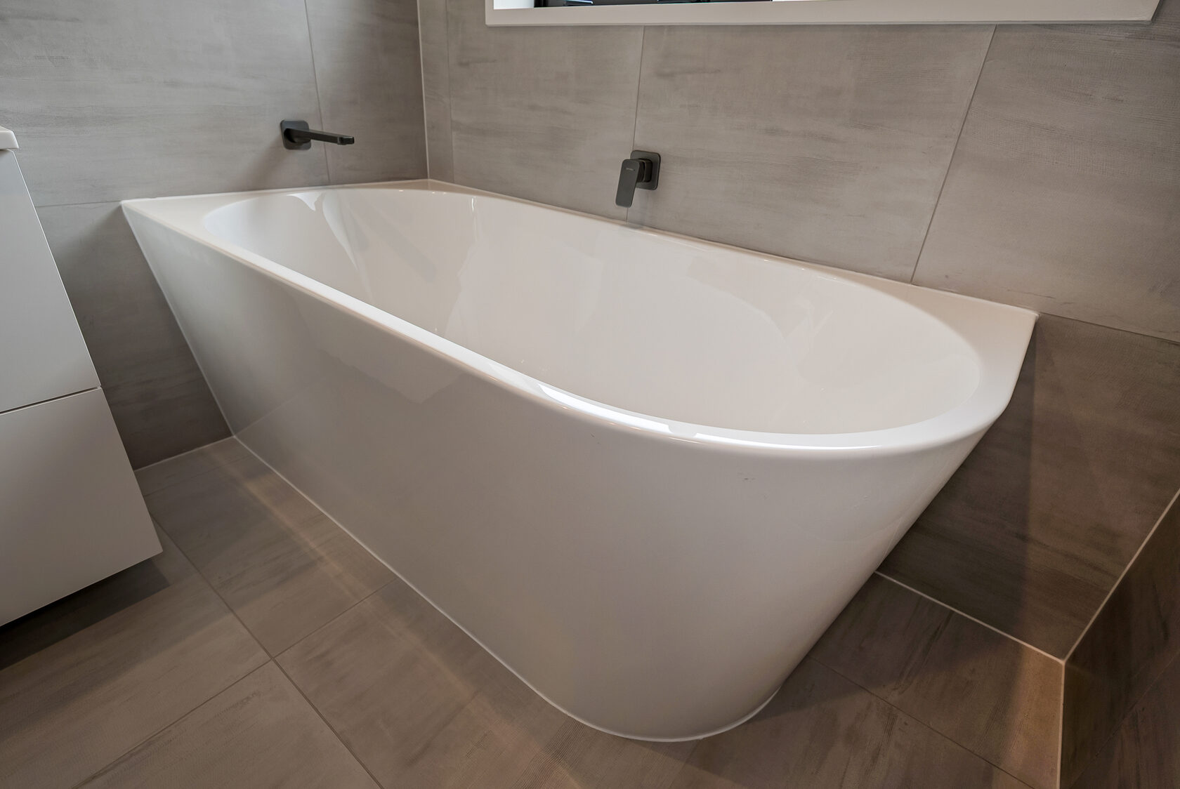 Modern freestanding bathtub with smooth curved edges, matte black wall-mounted bath spout and mixer, and large-format stone-look tiles for a clean, contemporary finish.
