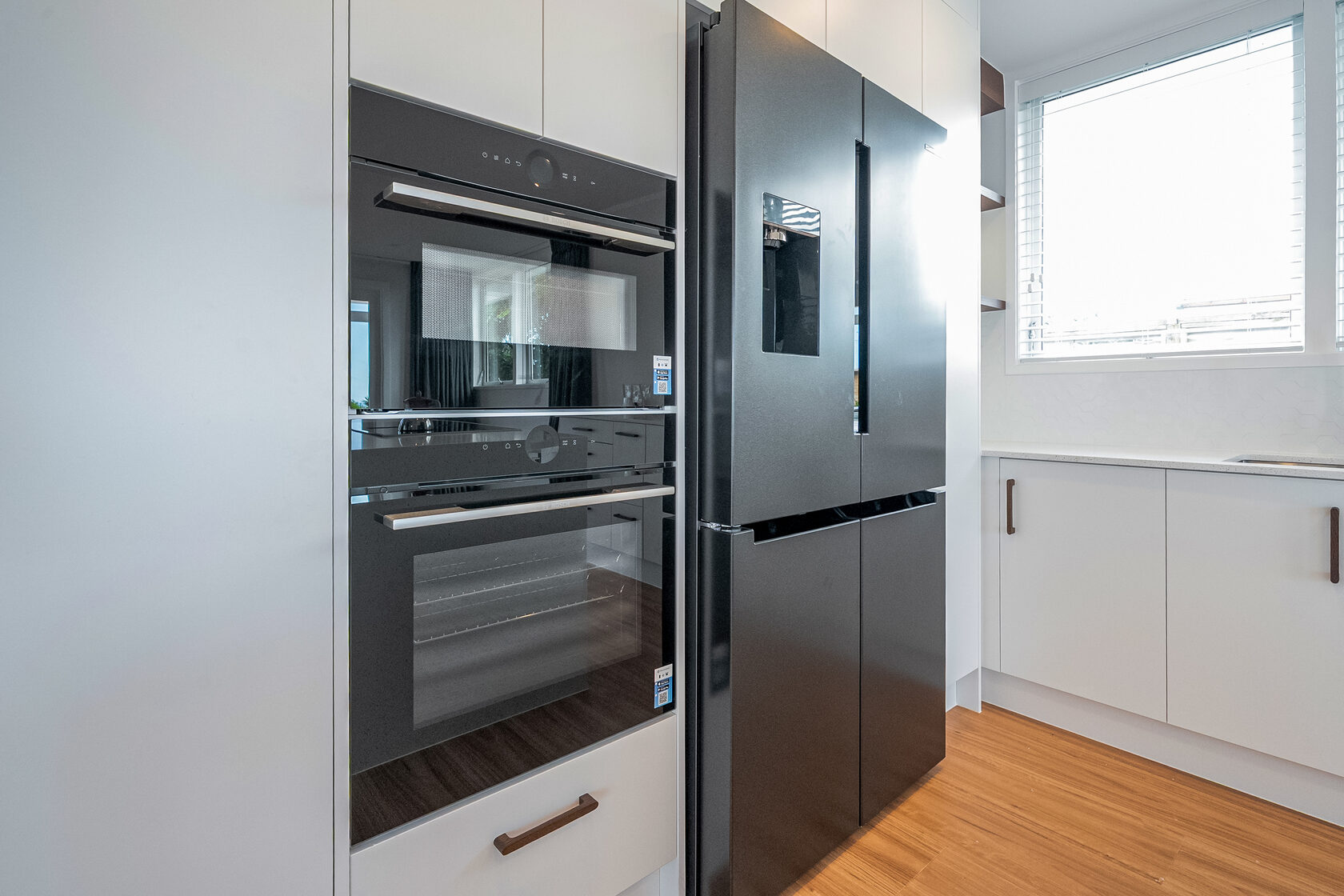 Modern kitchen with built-in black oven and double-door refrigerator set within white cabinetry and timber flooring.