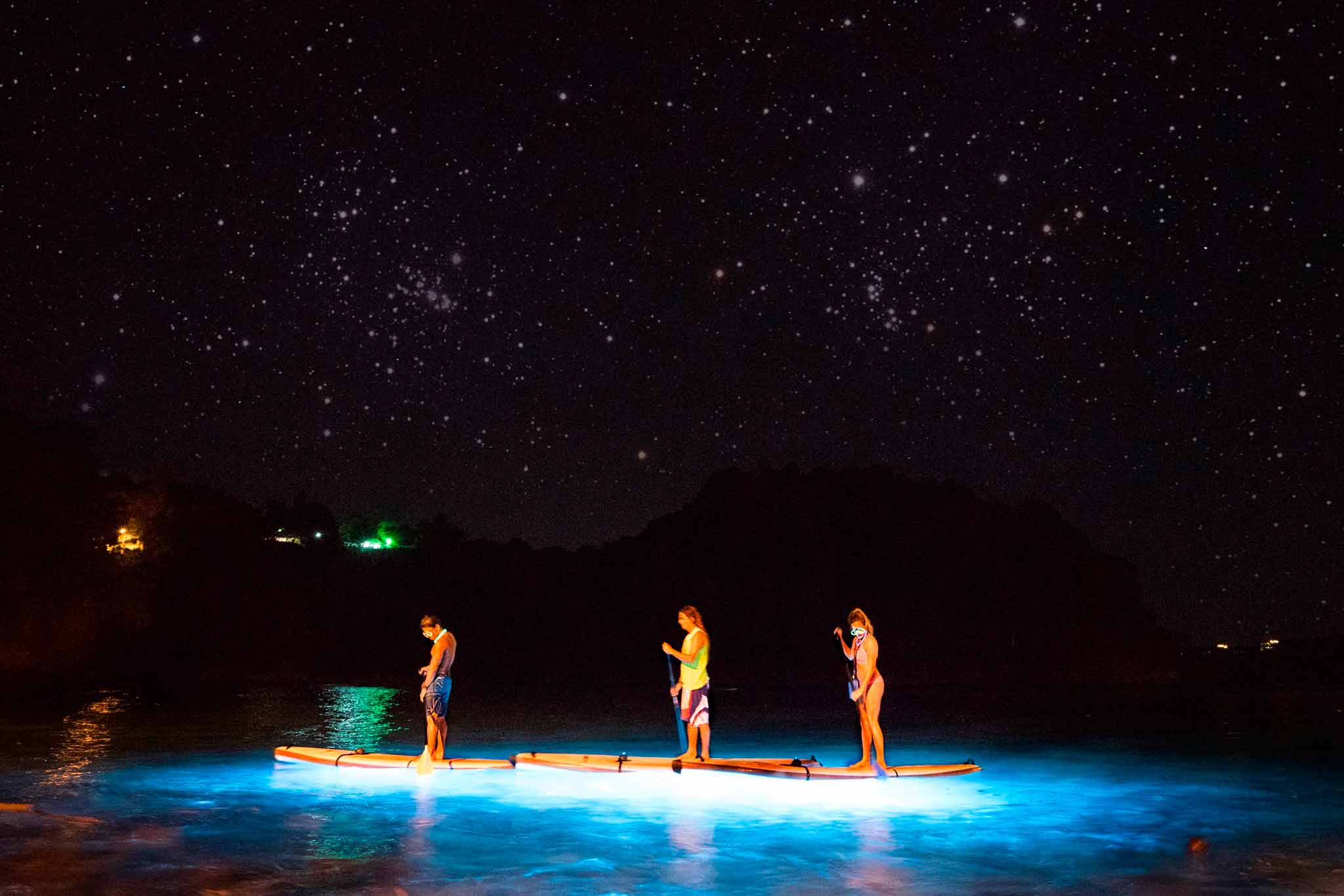 Tres personas sobre una tabla de paddle iluminada bajo un cielo estrellado