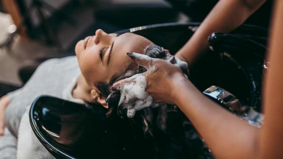 The image shows a woman having her hair washed in a salon, illustrating how hard water can impact hair health by causing dryness, buildup, and reduced lather formation