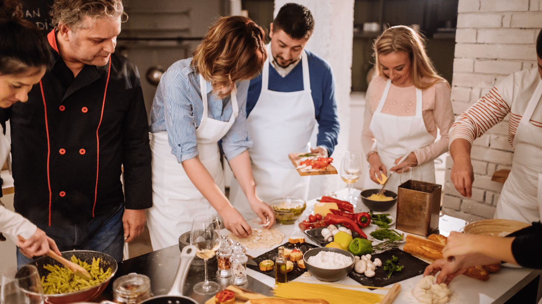 Gastronomic team building. A group of people, led by a professional chef in a black tunic, participates in a cooking class on making Italian pasta in a bright kitchen. A home-like atmosphere and fresh products on the table.