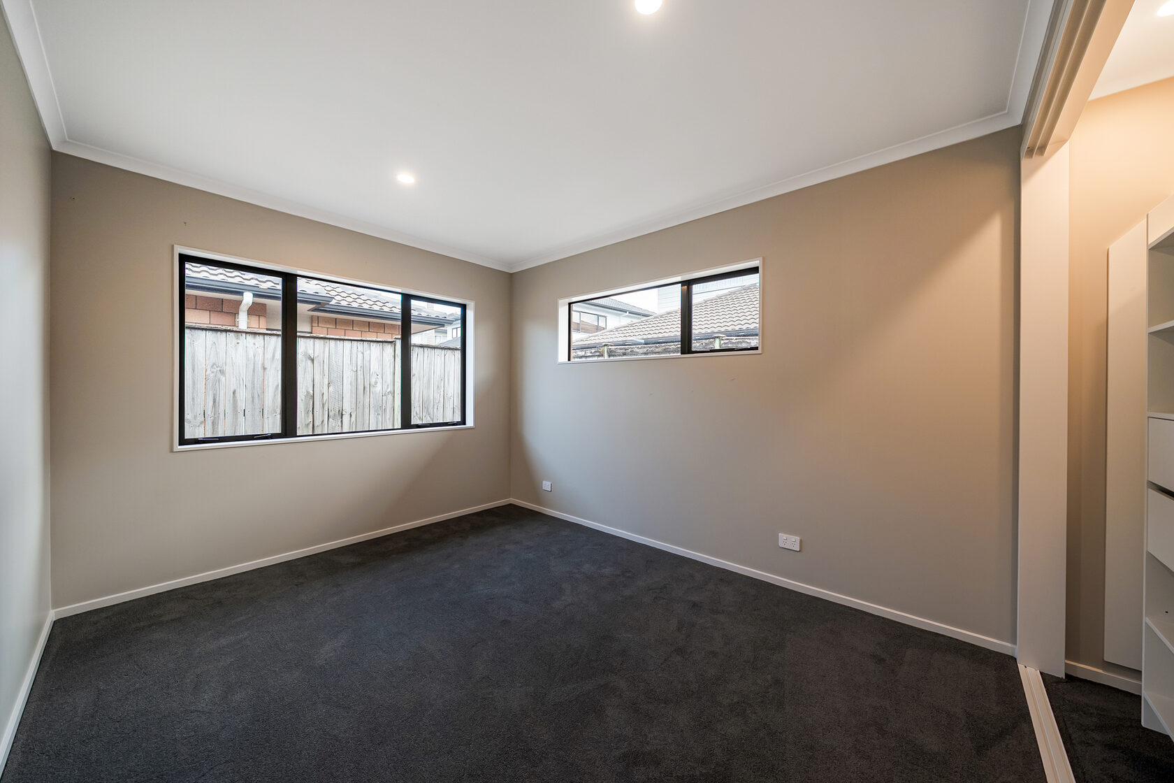 Empty carpeted bedroom with beige walls, recessed ceiling lights, and two black-framed windows facing a fenced backyard.