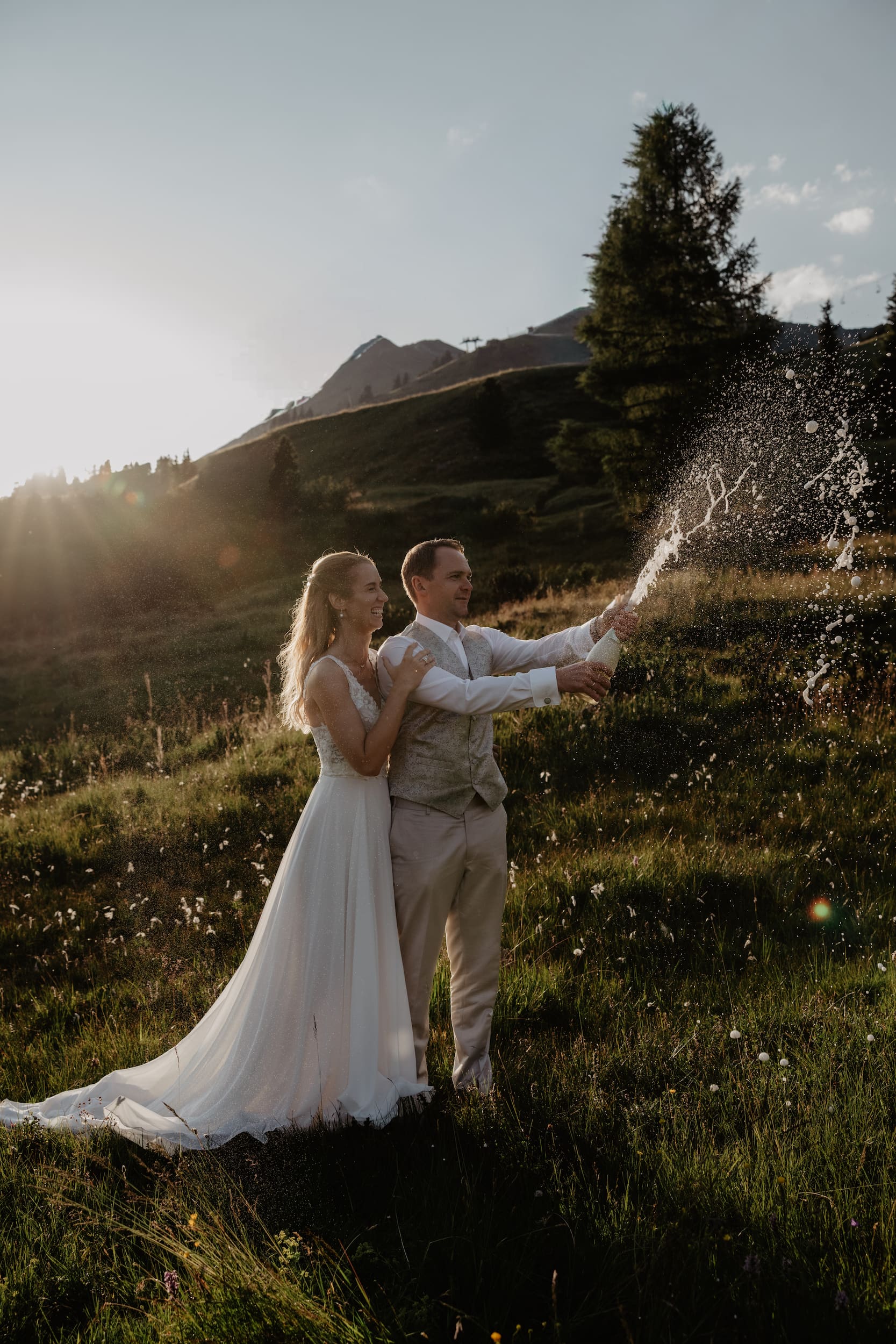 Newlyweds celebrating with a spray of champagne in a sunlit alpine meadow, surrounded by mountain scenery