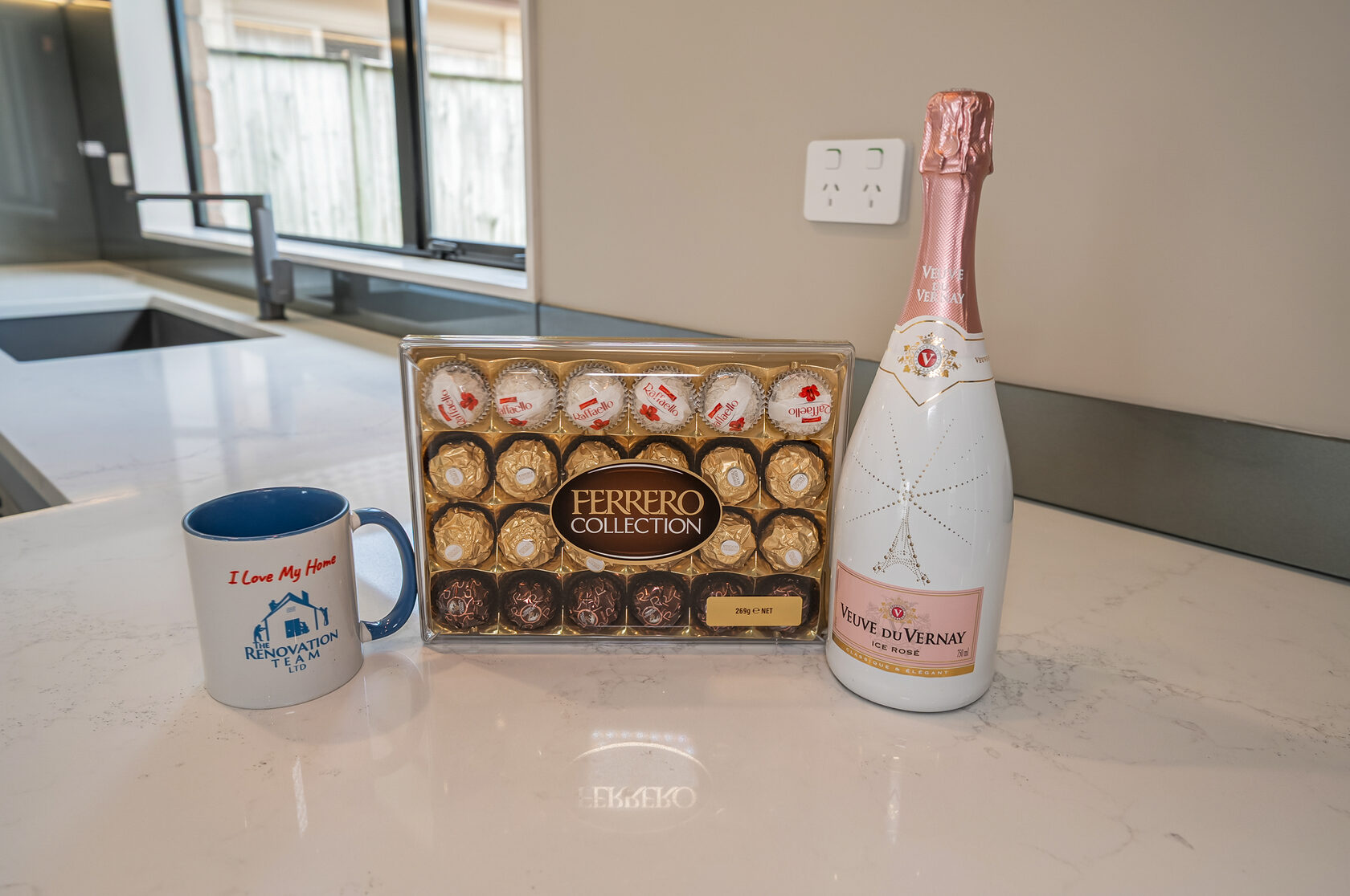 Box of assorted chocolates and a bottle of sparkling rosé placed on a kitchen countertop next to a branded coffee mug.