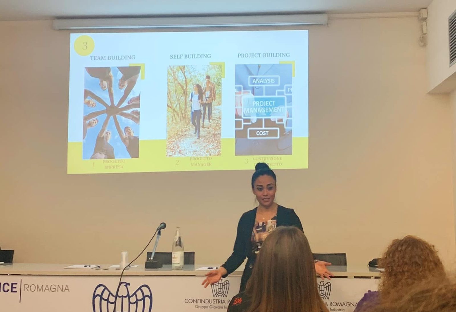 Ana Laura Ramirez in piedi davanti allo schermo mentre parla a una sala, al tavolo dei relatori il marchio di Confindustria Romagna