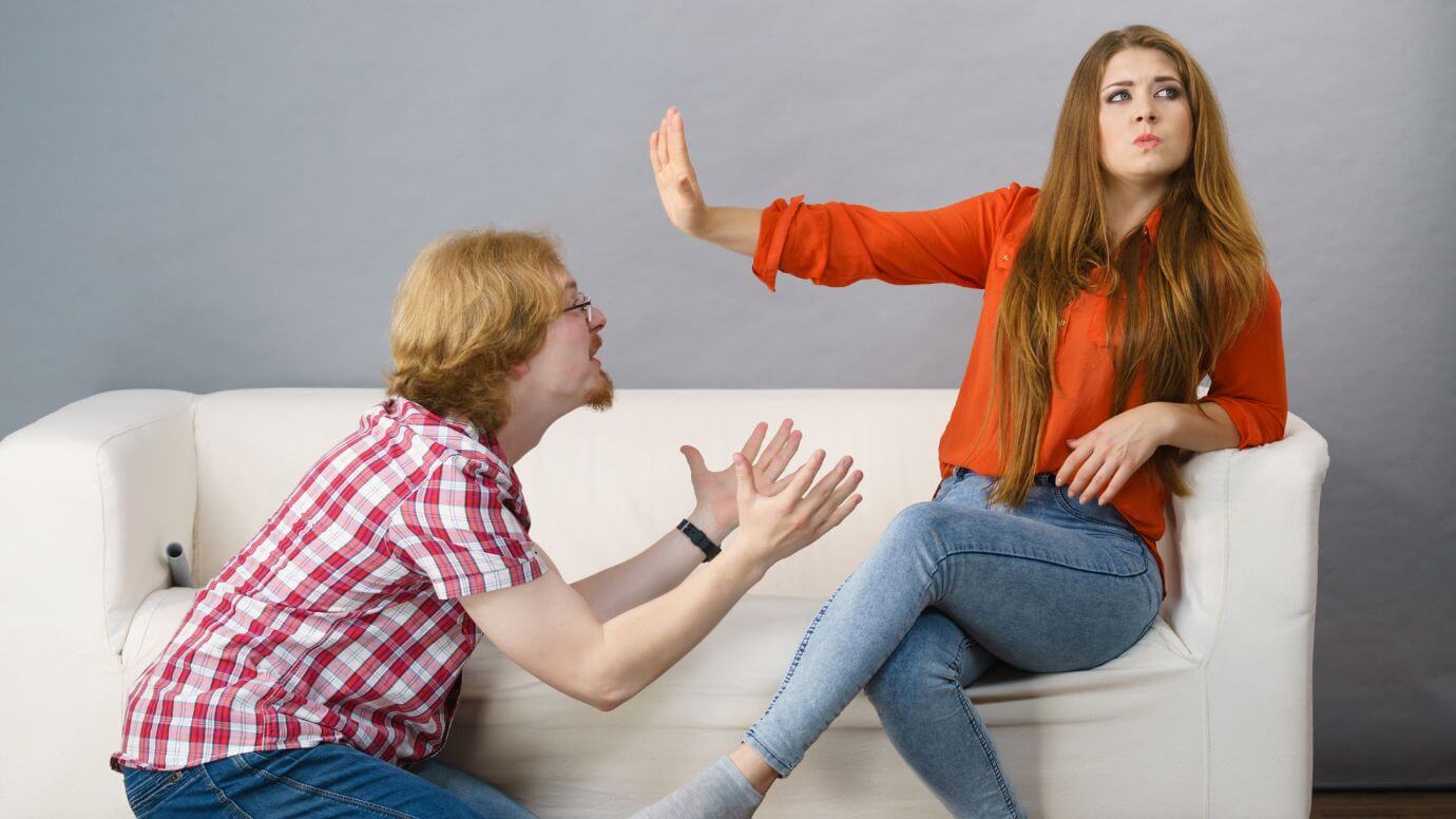 Redhead man humorously begging on his knees, while a redhead woman raises her hand in a gesture signaling him to halt