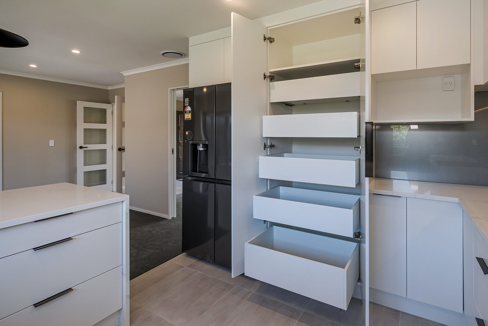 Modern kitchen with pull-out pantry drawers, white cabinetry, black double-door fridge, and sleek grey splashback.