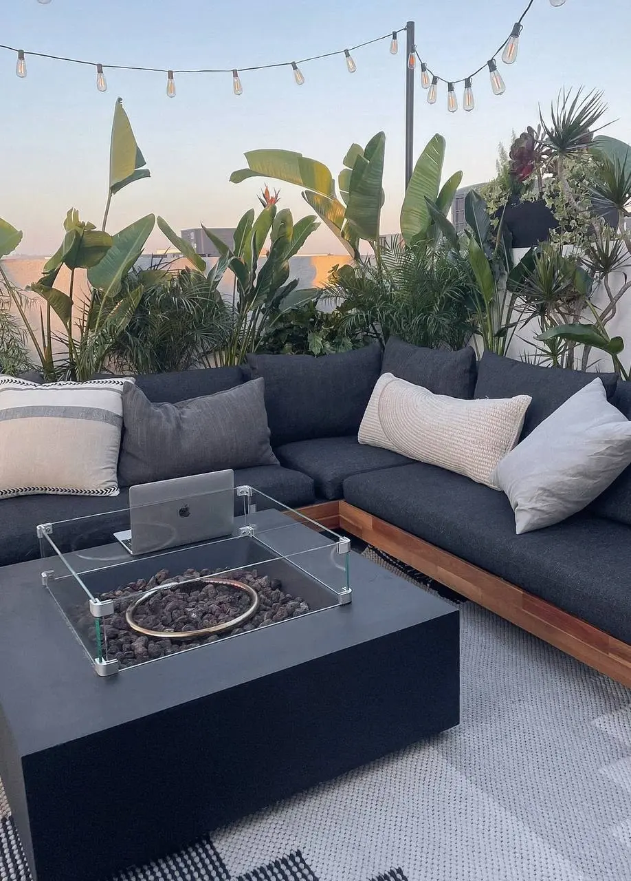 A rooftop lounge at dusk with a dark sectional and fire pit table, backed by bird of paradise and tropical planting against a city skyline