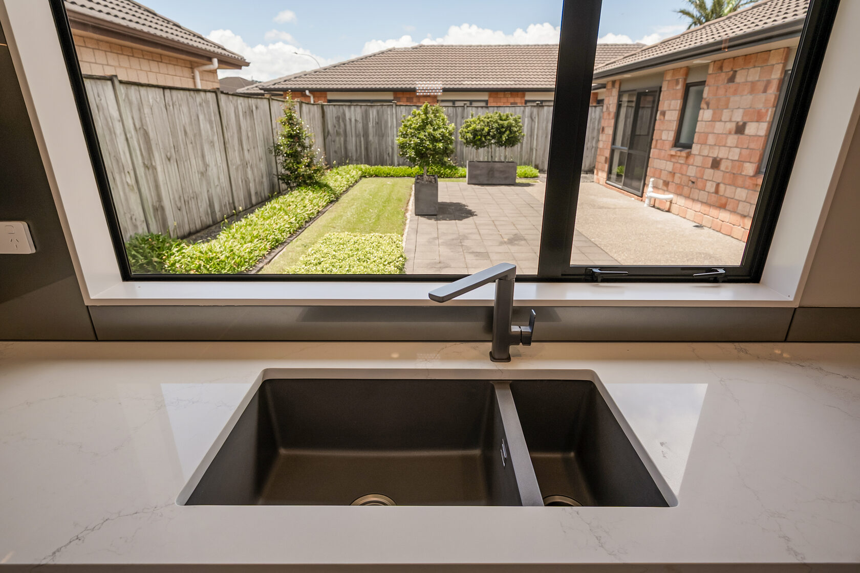 Double kitchen sink with modern black tap set into a stone countertop, overlooking a fenced backyard with lawn, patio, and brick exterior walls.
