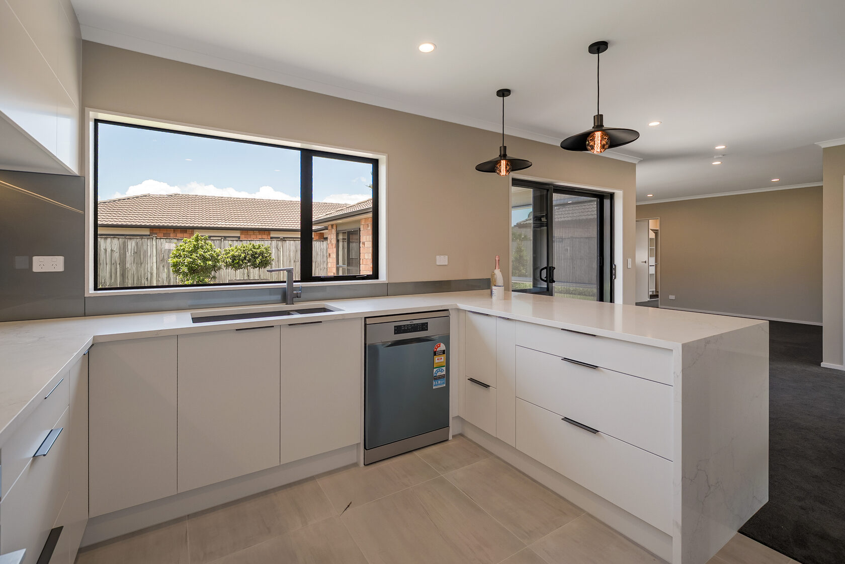 Bright modern kitchen with white cabinetry, L-shaped countertop, stainless steel dishwasher, large window overlooking the yard, and contemporary black pendant lights.