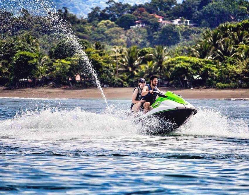 Dos personas en una moto acuática verde y blanca levantando espuma cerca de una playa tropical
