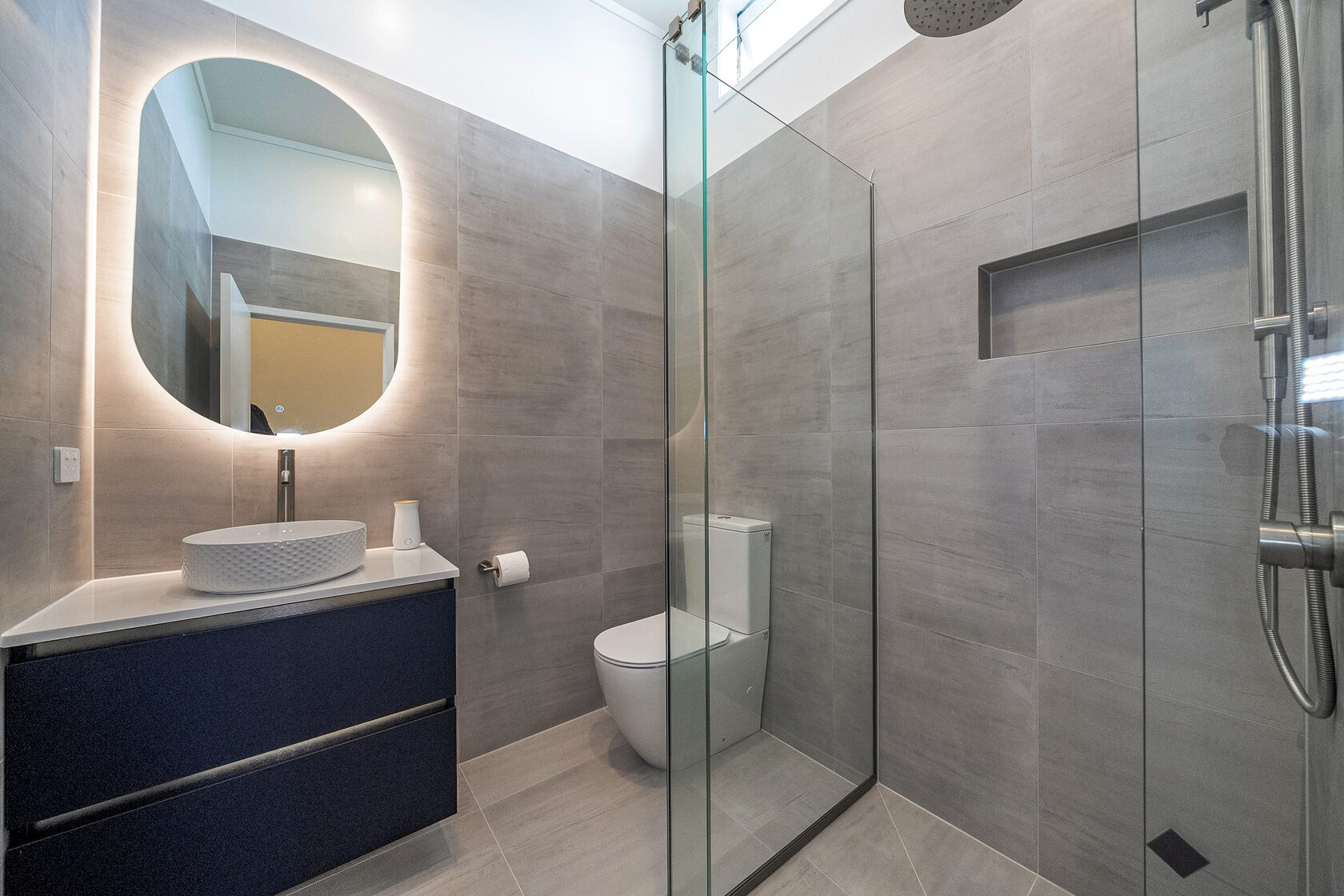 Modern ensuite bathroom with glass walk-in shower, grey tiled walls, floating vanity, and backlit mirror.