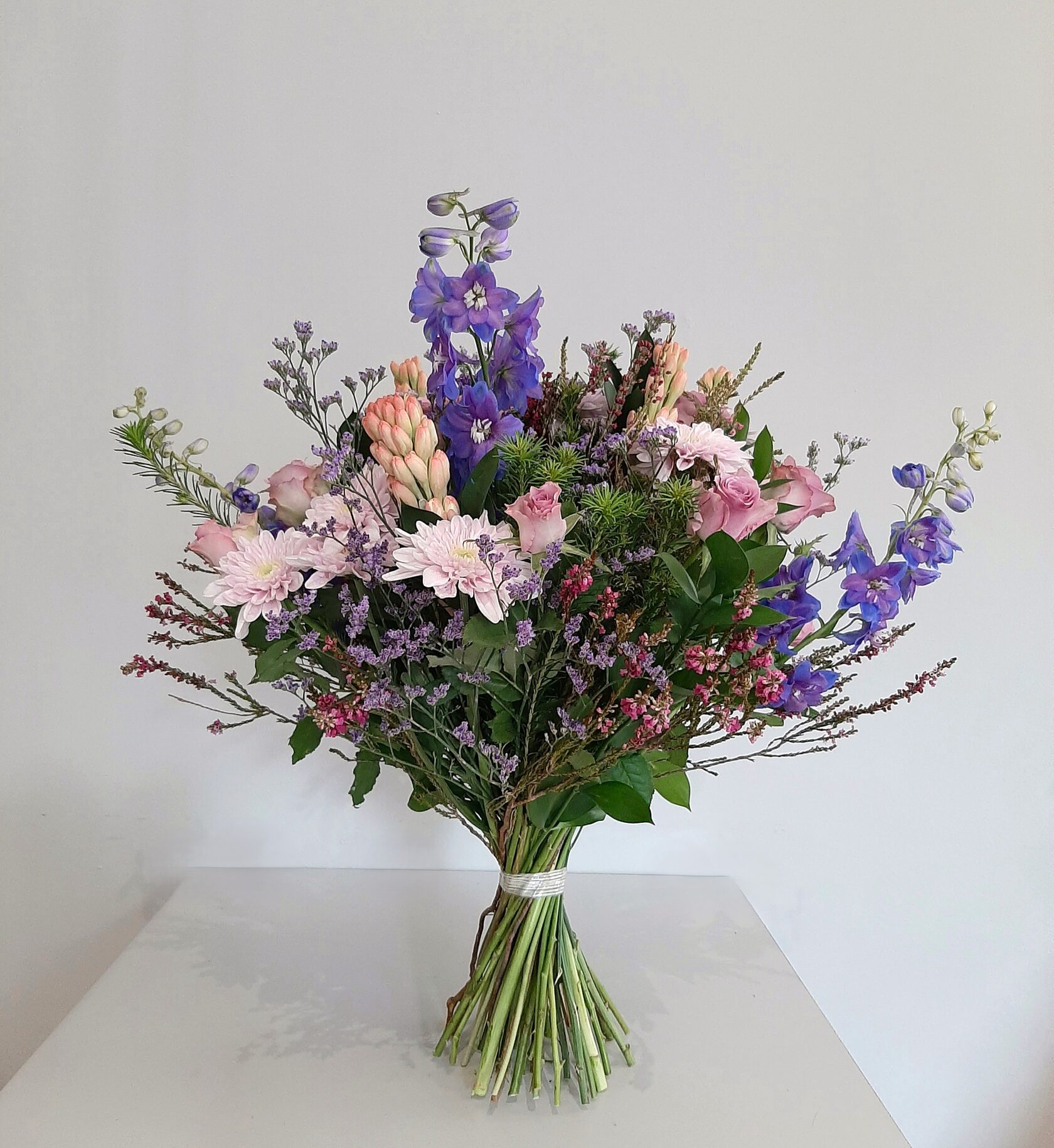 A lush garden bouquet featuring delphiniums, pink chrysanthemums, roses, and hyacinths in a delicate white, pink, and purple color palette.