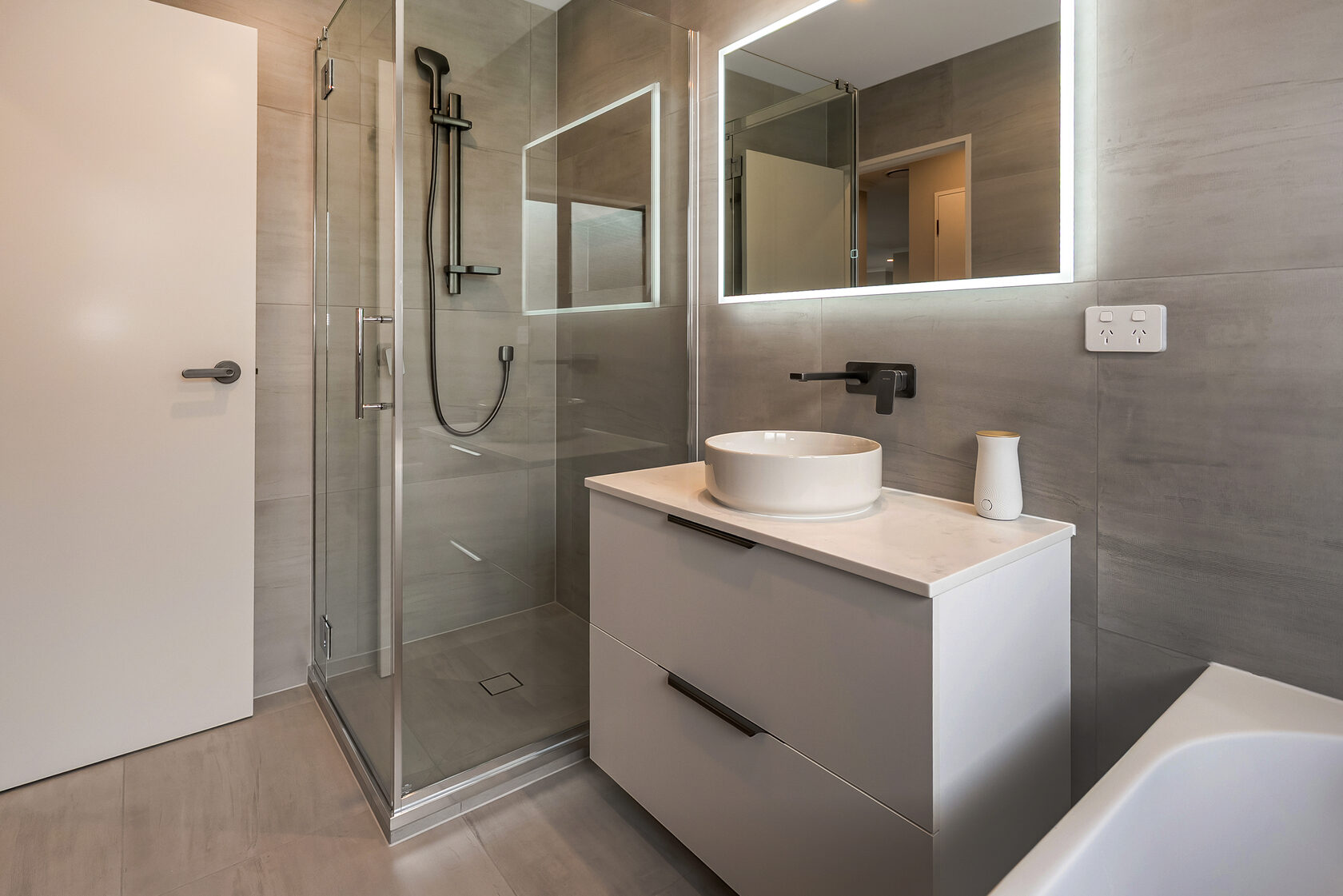 Contemporary bathroom featuring a glass walk-in shower with matte black fixtures, floating white vanity with round vessel basin, wall-mounted tapware, and LED backlit mirror against large grey tiles.