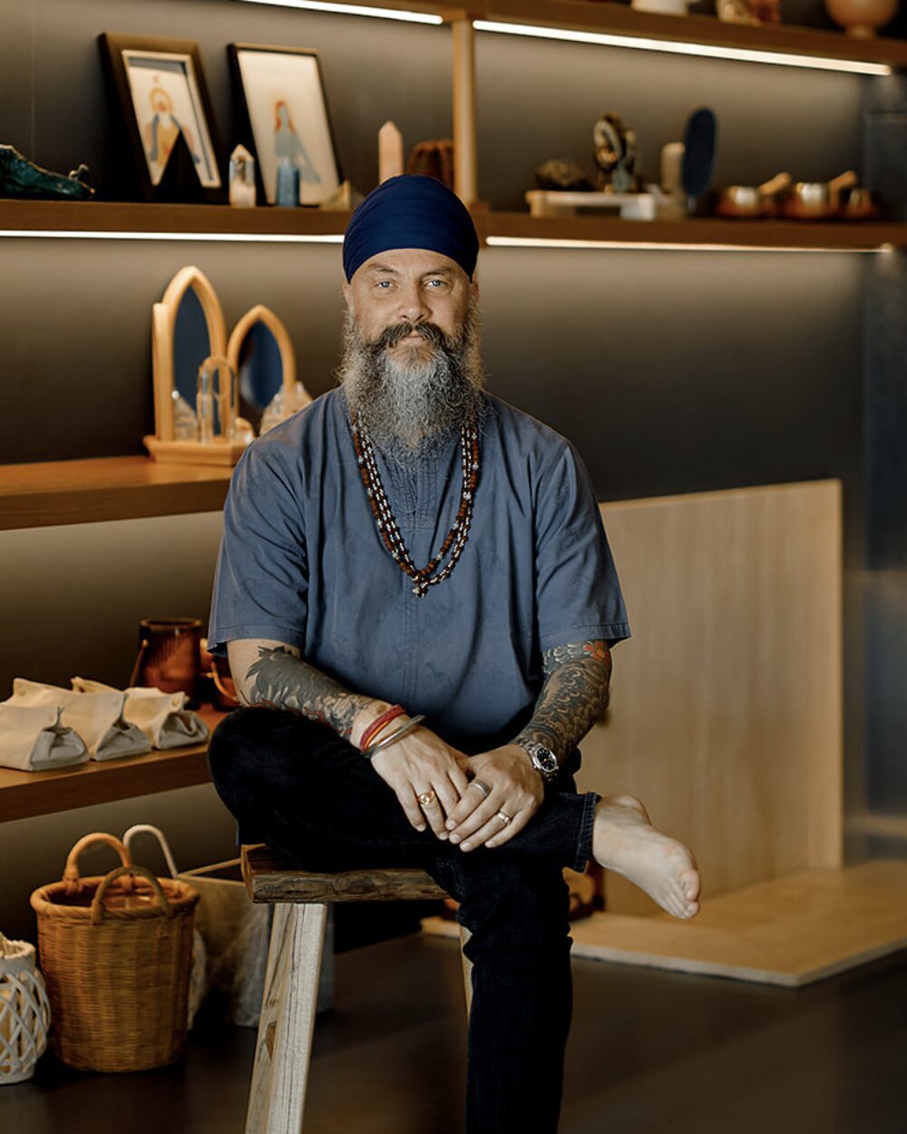 Jugat Guru in a blue turban and shirt, seated in a warm-lit room with shelves of singing bowls and crystals