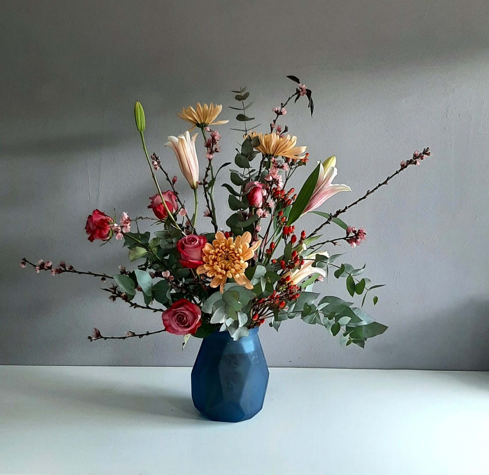 A floral arrangement of roses, chrysanthemums, lilies, and eucalyptus in a blue faceted vase against a light background.