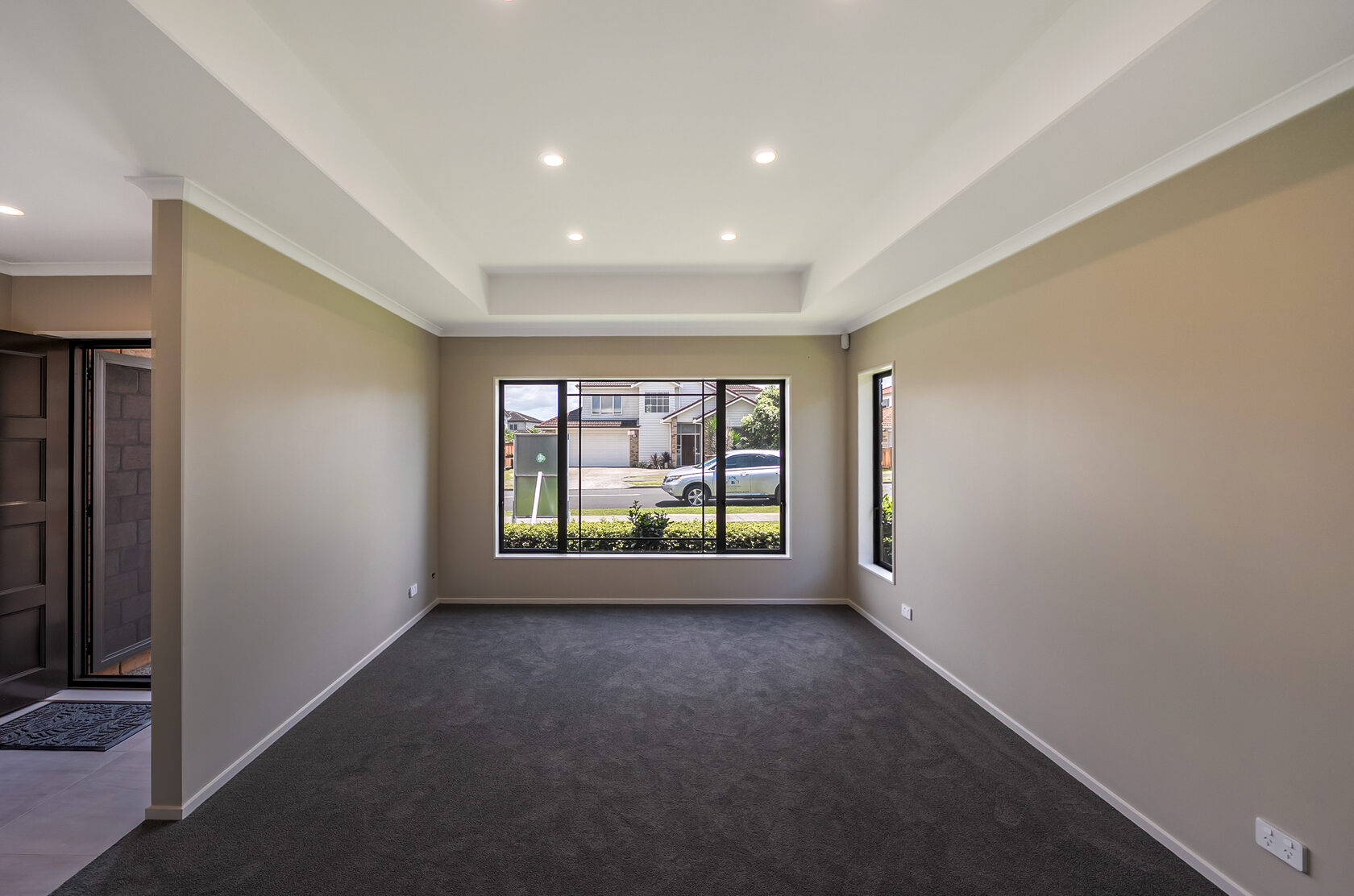 Flat Bush home renovation: Empty living room with recessed ceiling, downlights, neutral walls, dark carpet, and large front-facing window allowing natural light.