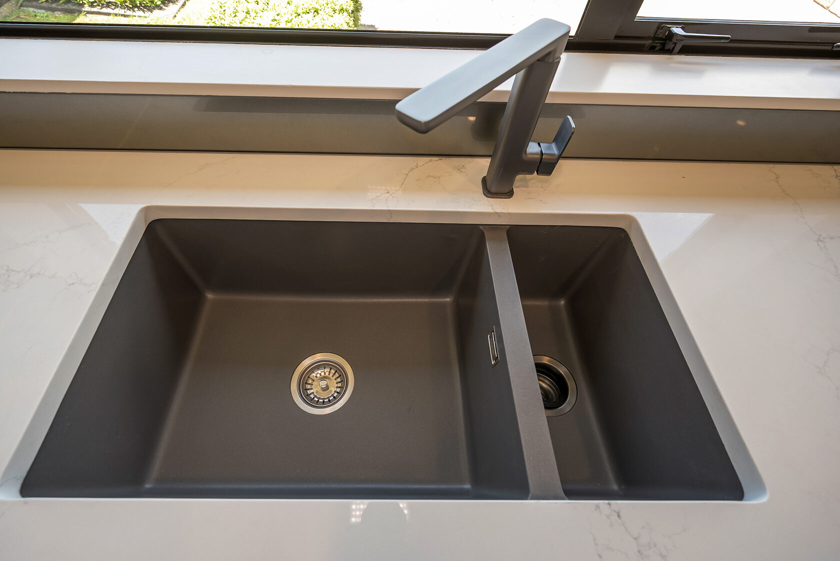 Close-up of modern double-bowl kitchen sink with matte black finish and matching angular tap set into a light stone countertop.