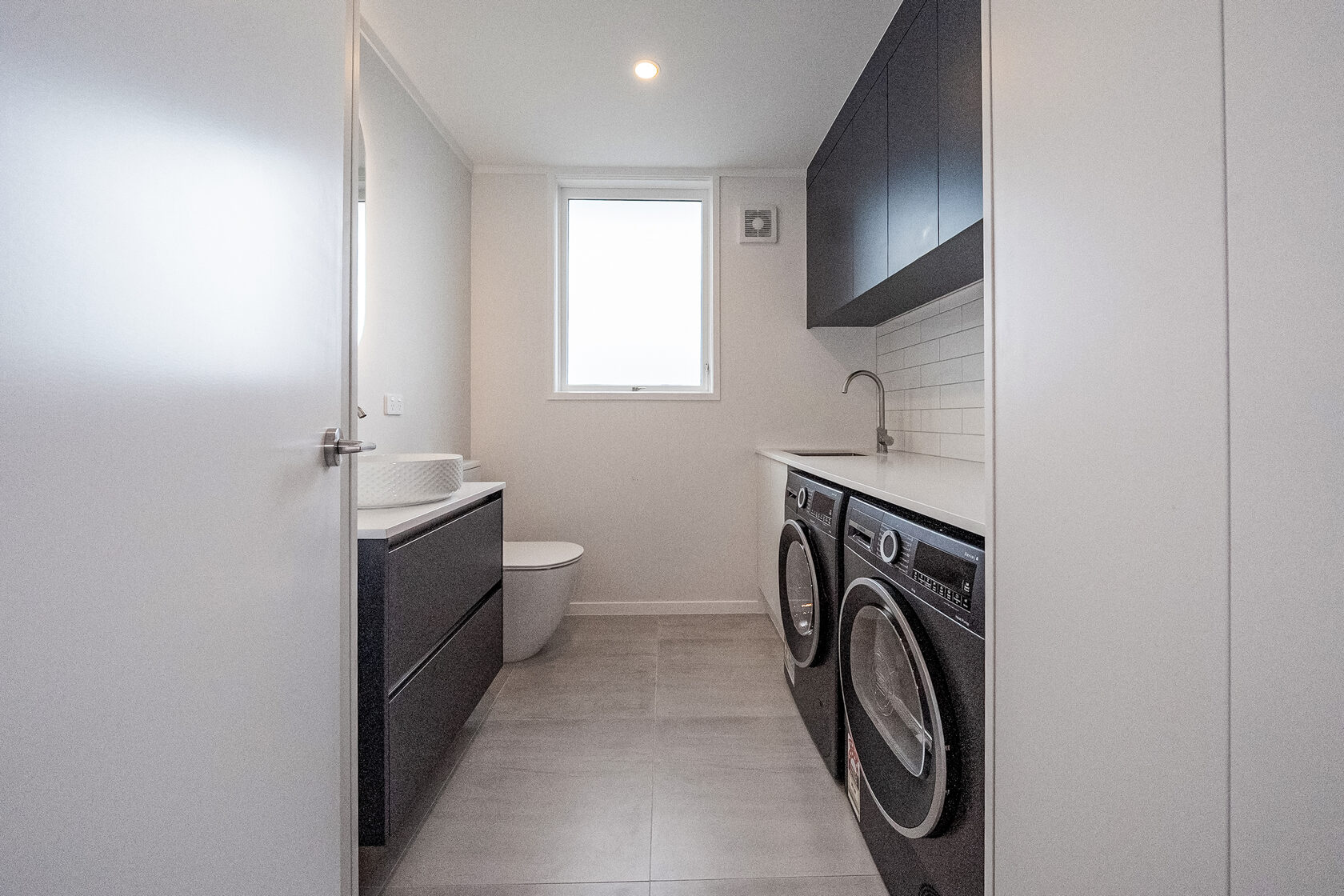 Compact modern bathroom and laundry combo with double front-loading washers, floating vanity, and tiled splashback.